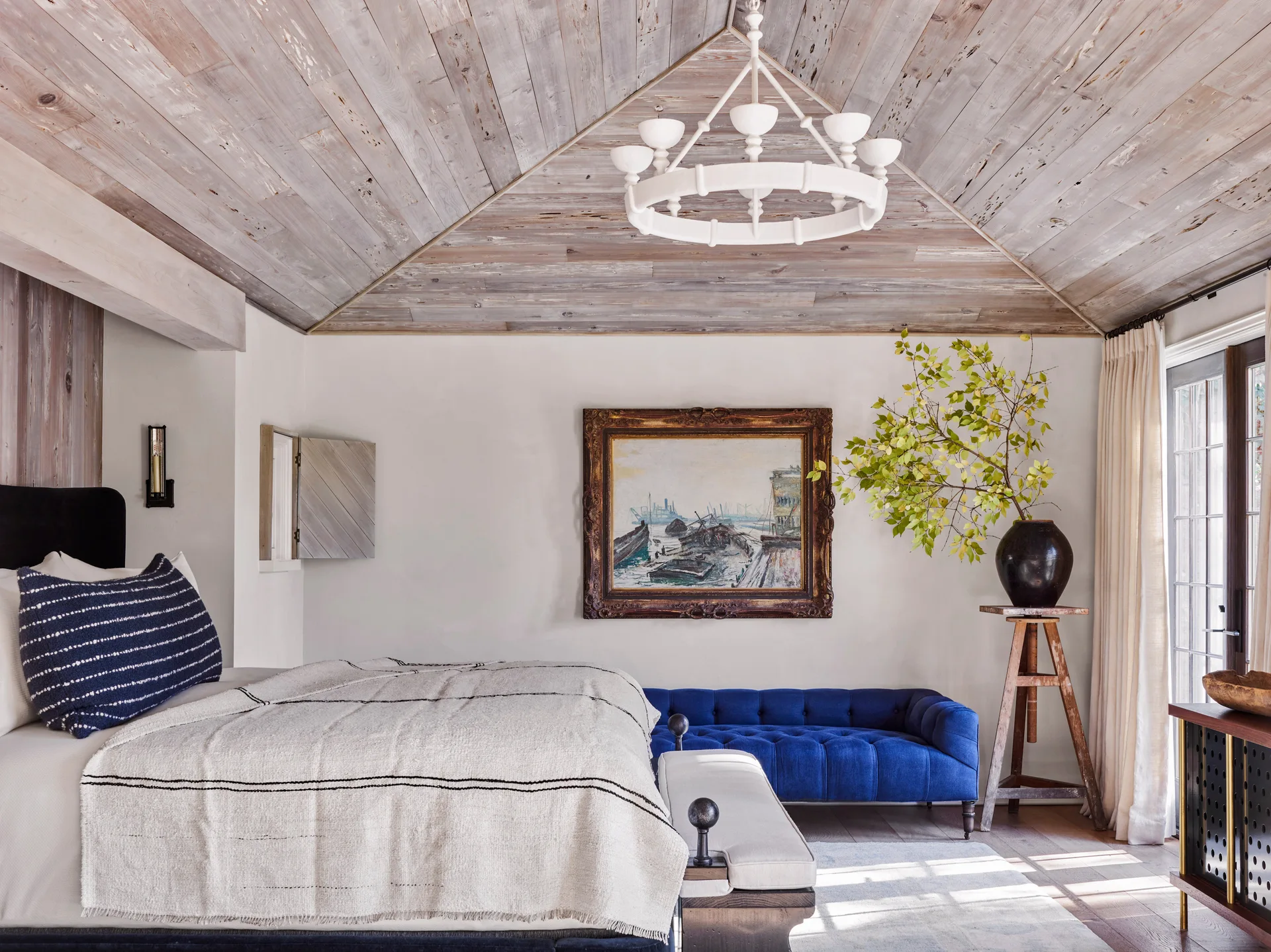 A bedroom featuring a vaulted whitewashed wood ceiling, a bed with textured linens, and a bright blue tufted velvet bench against the back wall.