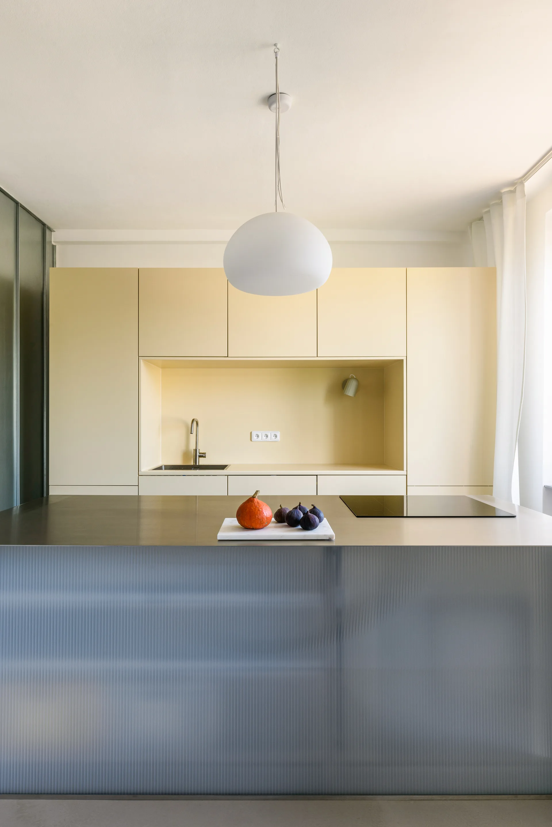 A modern kitchen features pale yellow built-in cabinetry, a metallic island with a fluted glass base, and a white globe pendant light.