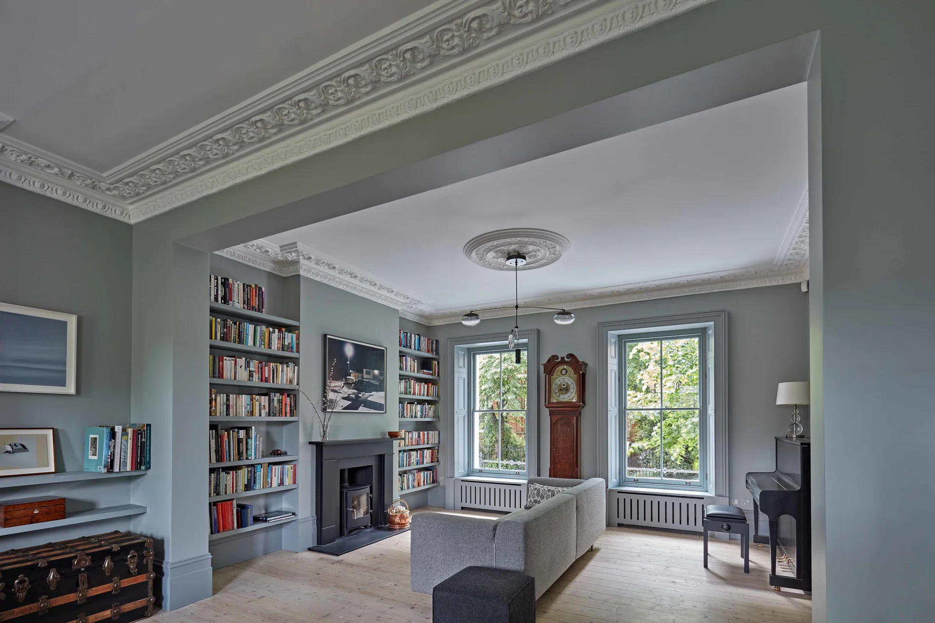 A grey living room featuring a central sofa, built-in bookshelves flanking a fireplace, ornate plaster crown molding, and large sash windows.