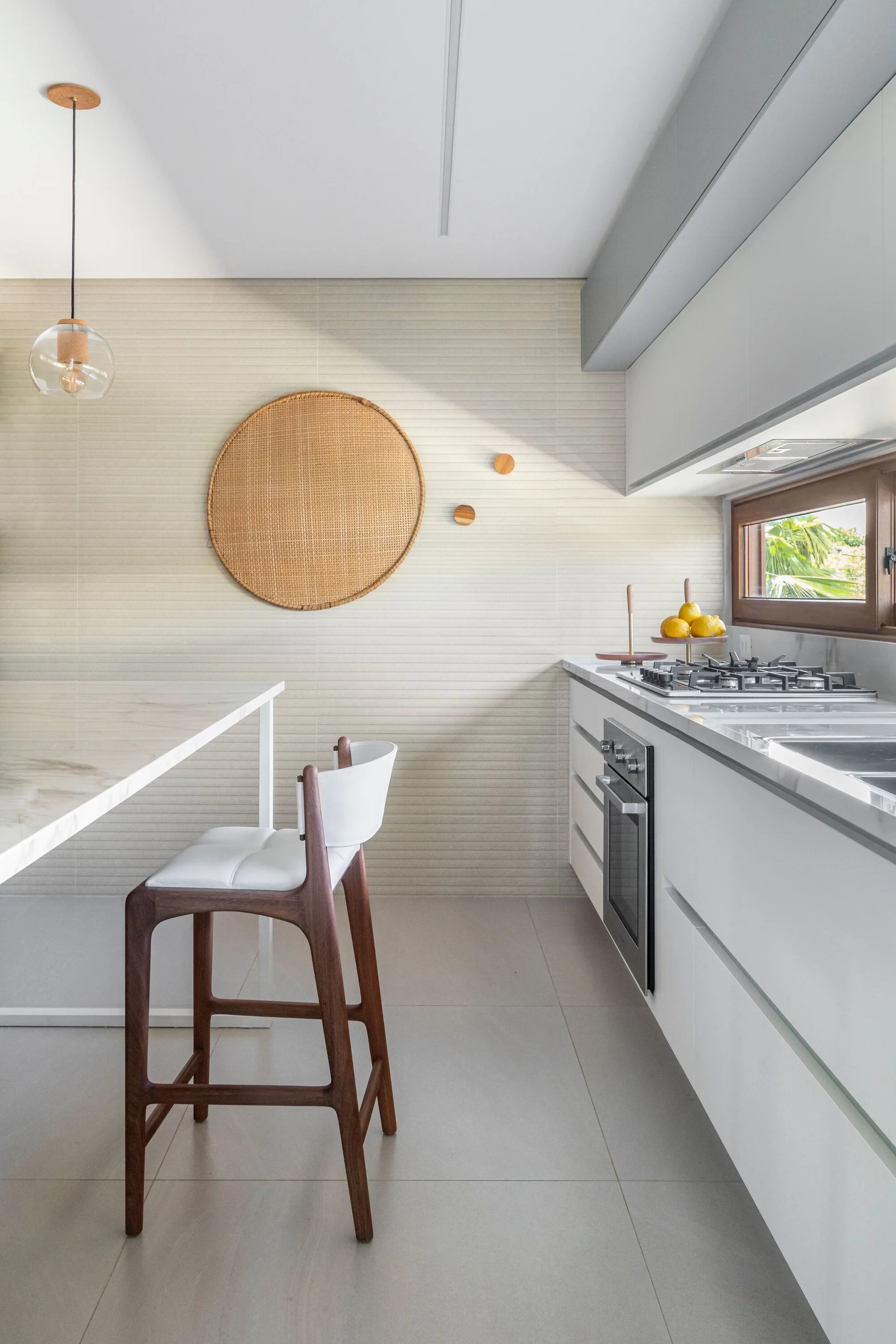 A minimalist white kitchen featuring a textured beige tile wall decorated with a round wicker ornament, a wooden bar stool, and white cabinetry.