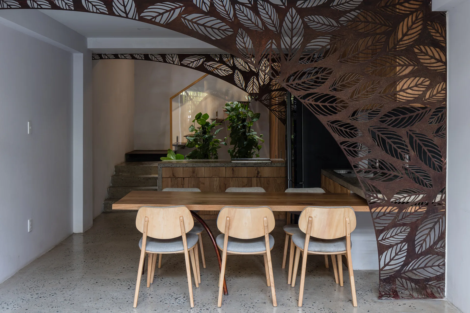 A wooden dining table and matching chairs stand on a speckled grey floor next to a rust-colored metal screen with a laser-cut leaf pattern.