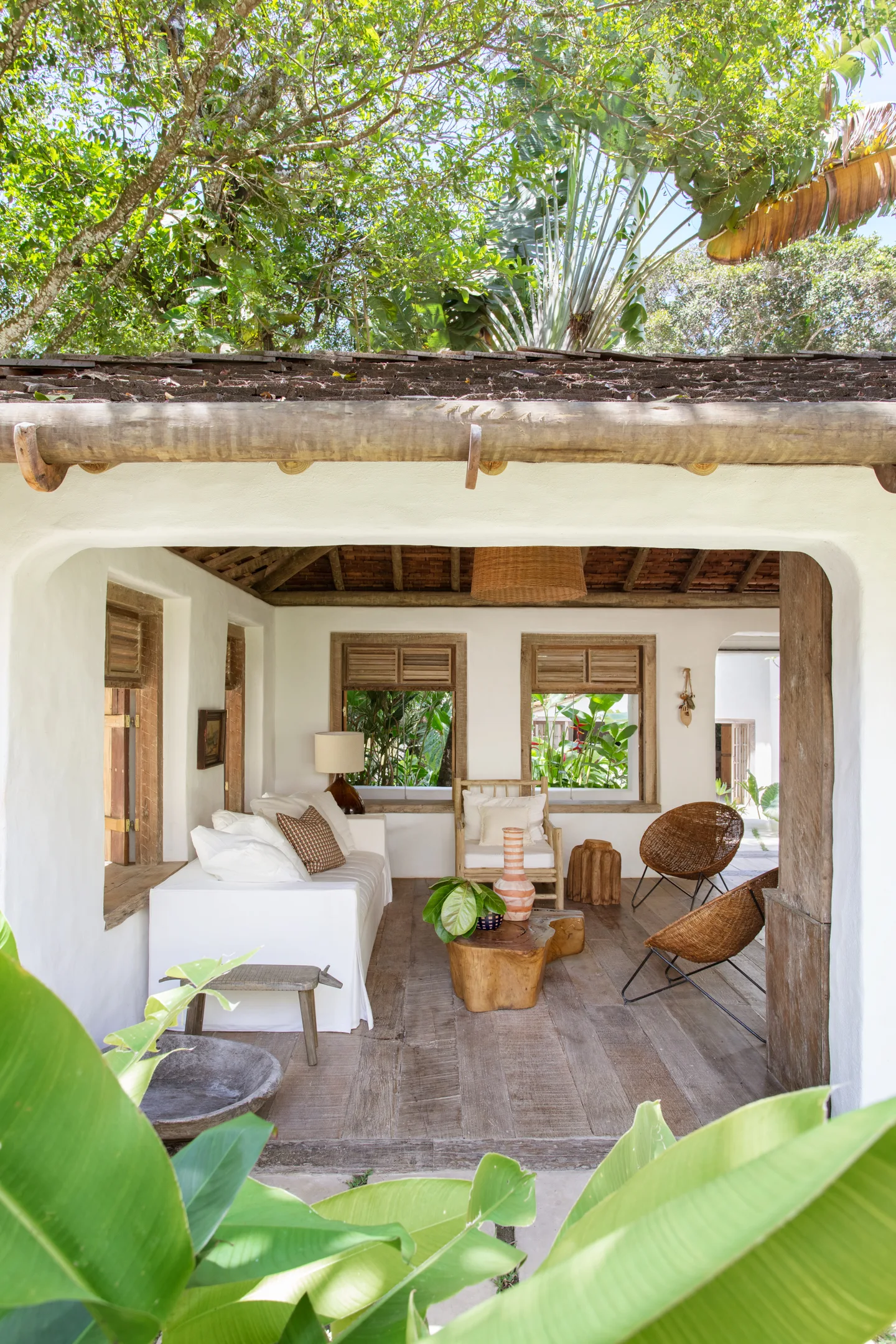 A covered rustic patio features a white sofa, woven wicker lounge chairs, a live-edge wood coffee table, and plaster walls framed by green trees.