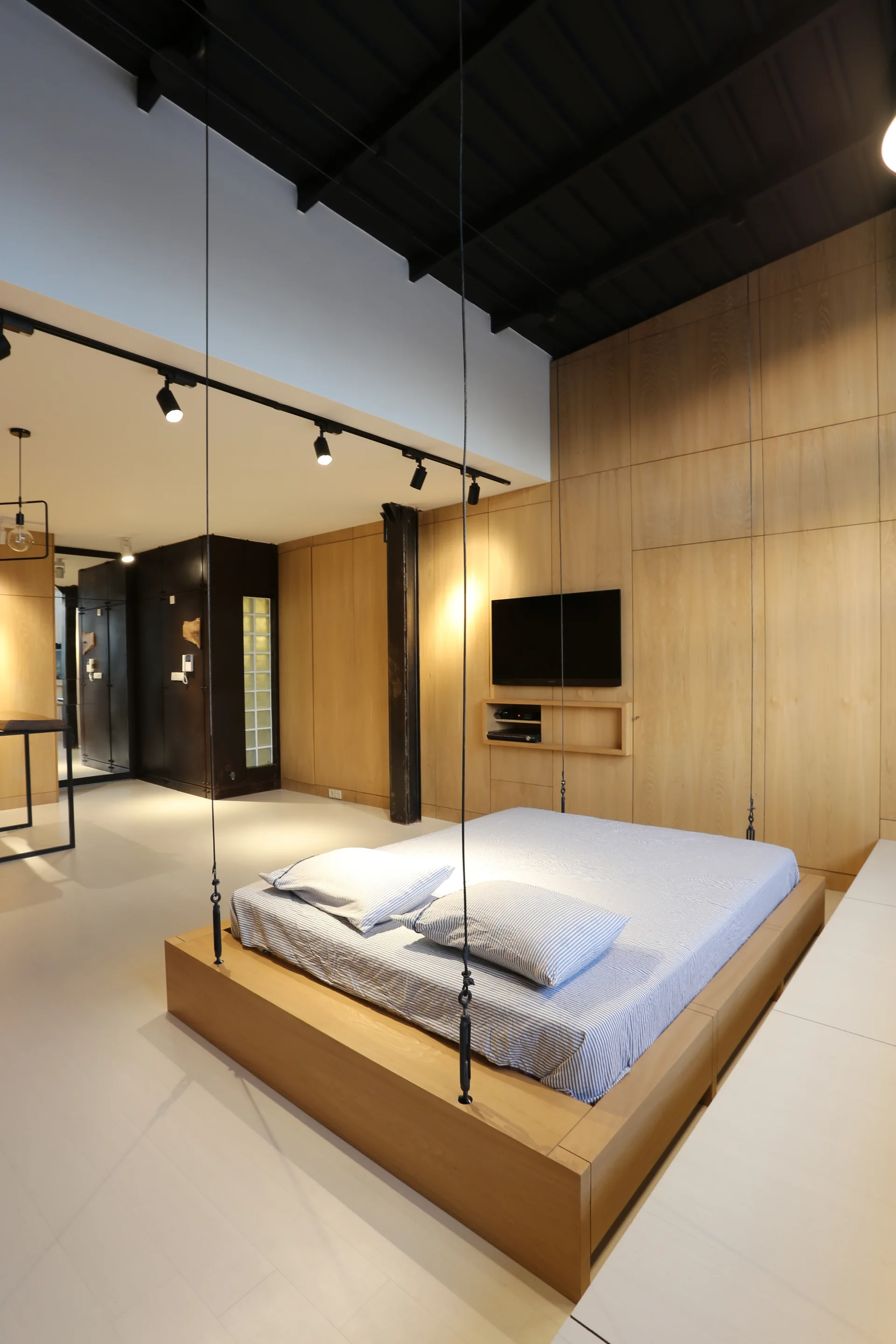 A modern industrial bedroom features a wood platform bed suspended by black cables in front of a light wood paneled wall.