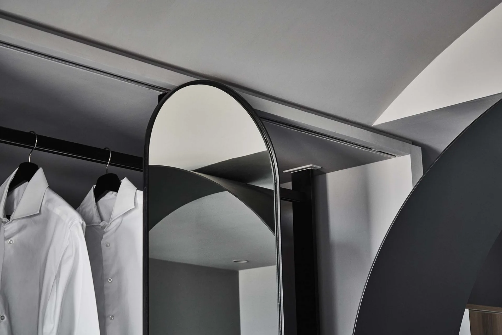 A minimalist grey closet features white shirts hanging on a black metal rail next to a sleek arched mirror.
