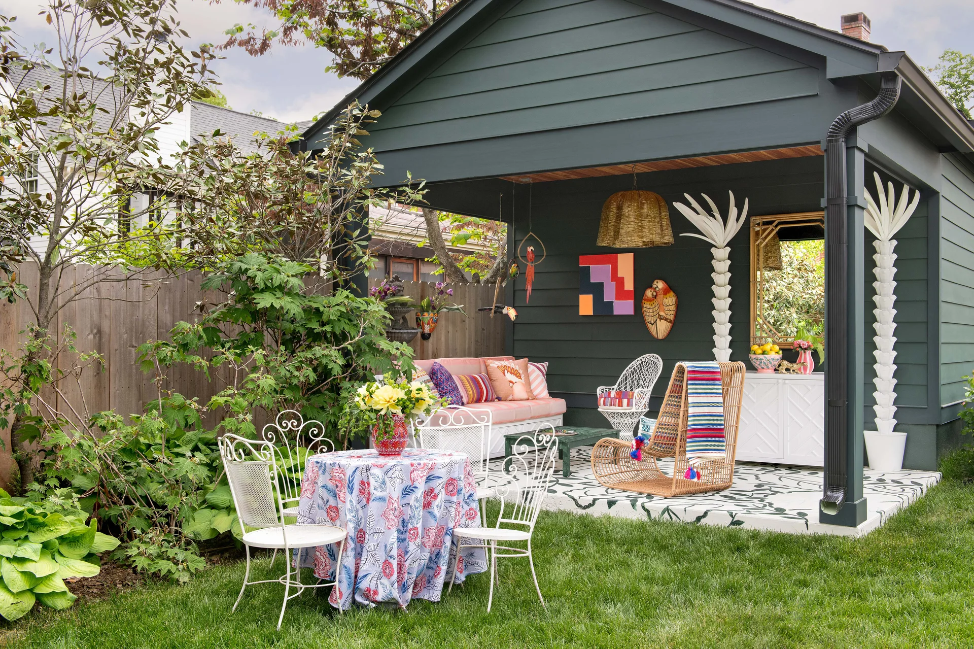 A covered backyard patio featuring dark green siding, white palm tree columns, eclectic furniture, and a painted botanical floor.
