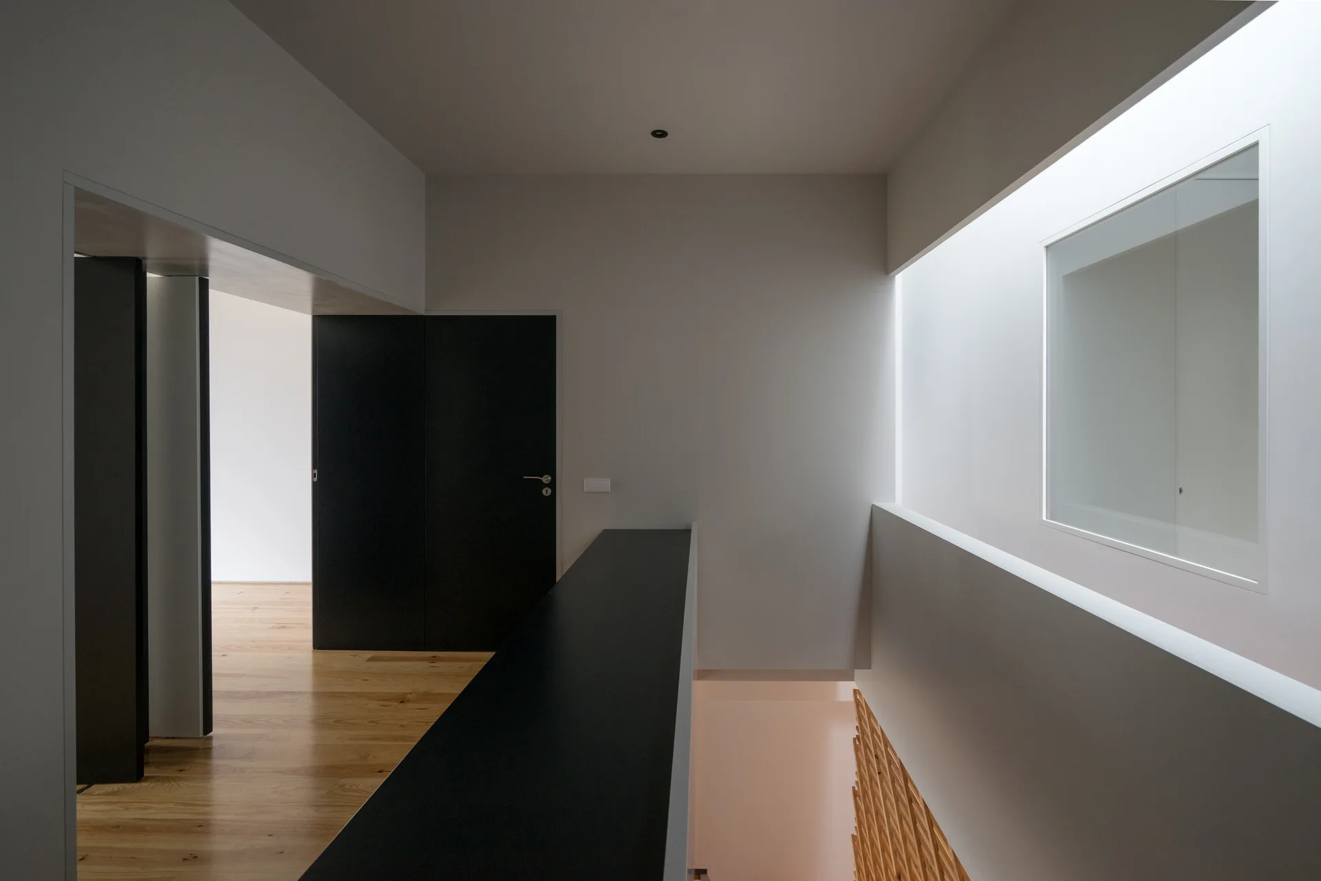 A minimalist stairwell landing features light wood flooring, a stark matte black door, and soft light from a deep overhead skylight.