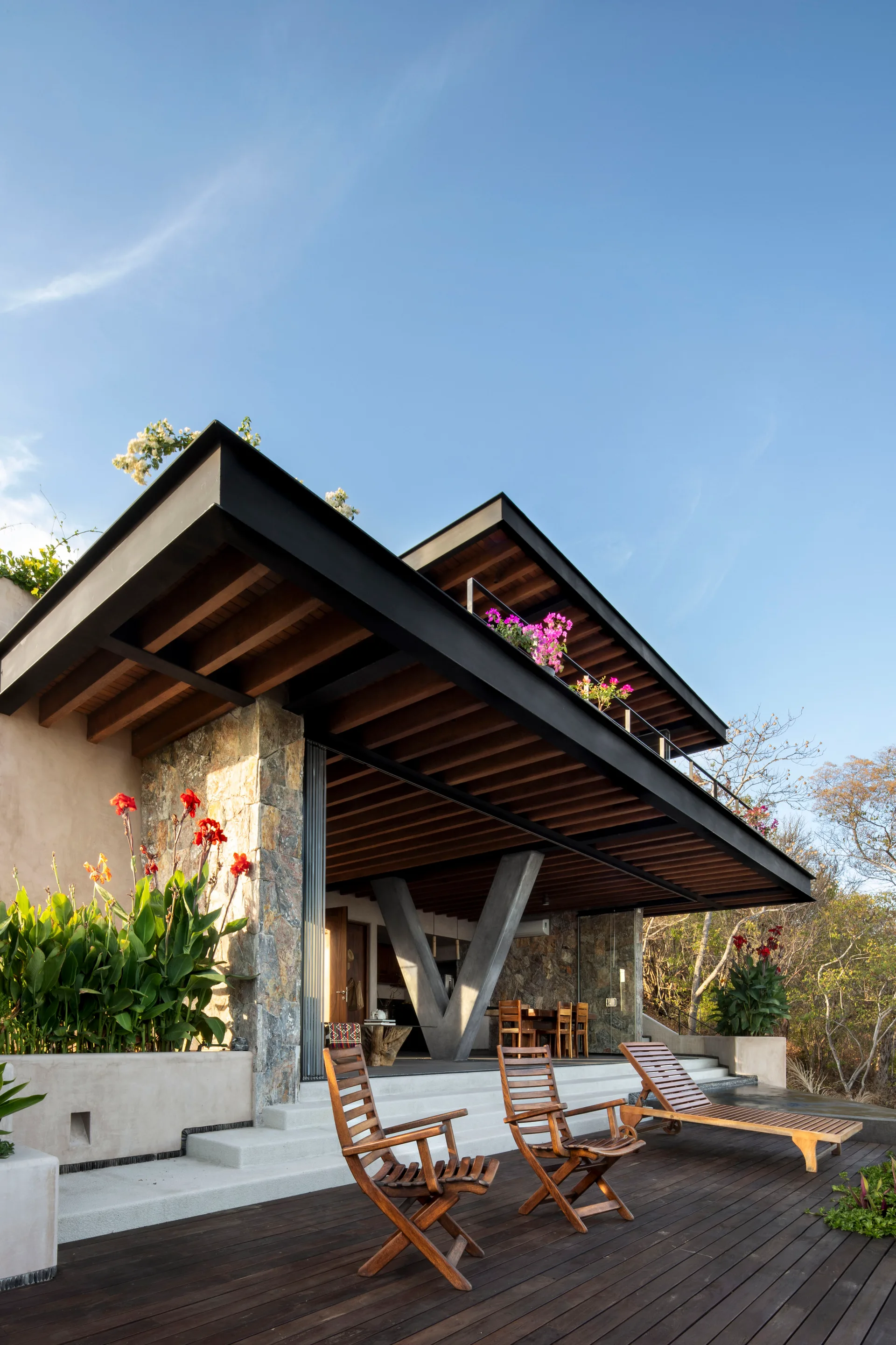 An outdoor wooden deck features matching timber lounge chairs and concrete steps leading to a covered patio with a V-shaped support column.