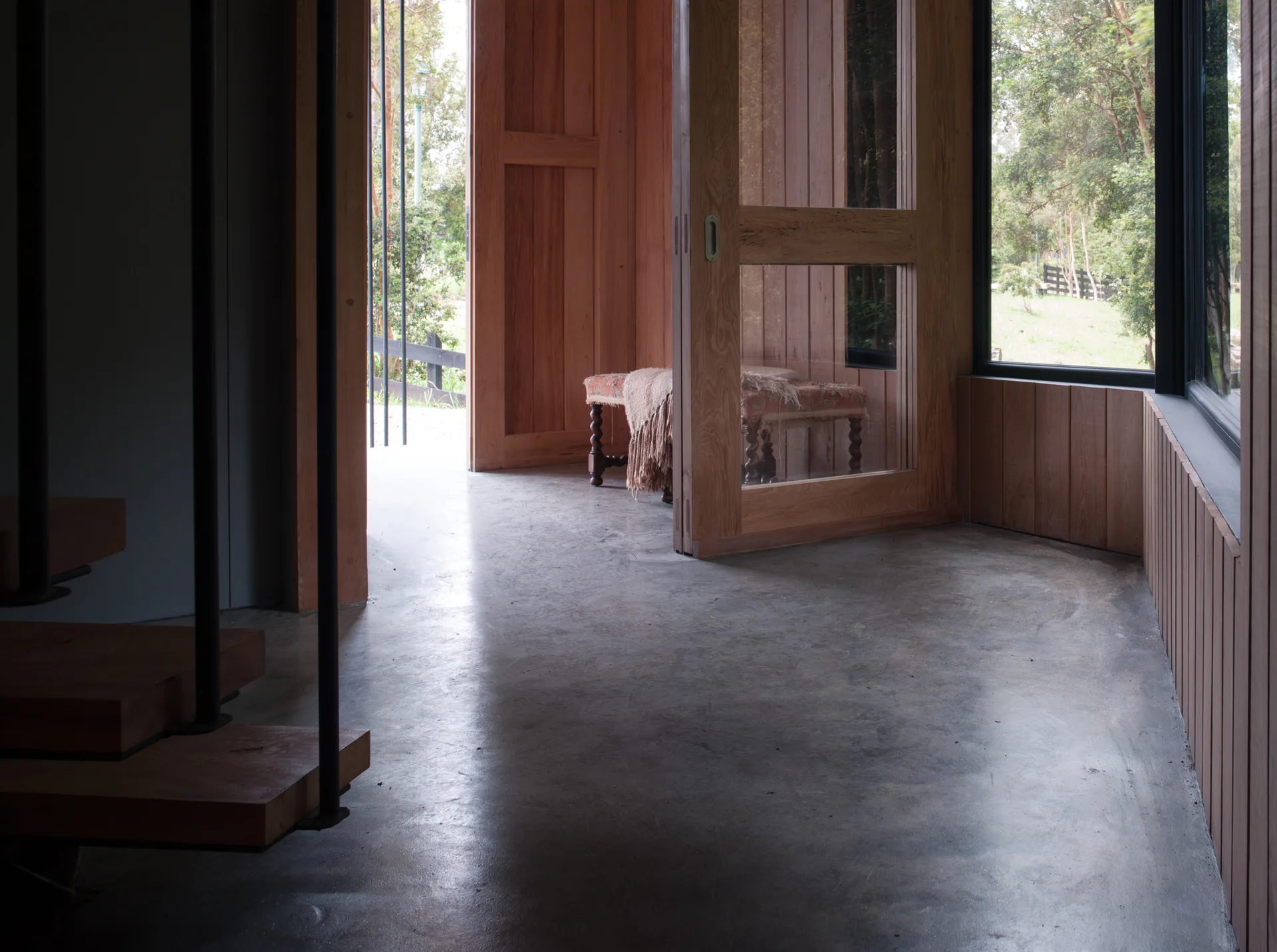 A rustic modern entryway featuring a polished concrete floor, vertical wood wall paneling, a wooden glass door, and wooden stairs on the left.