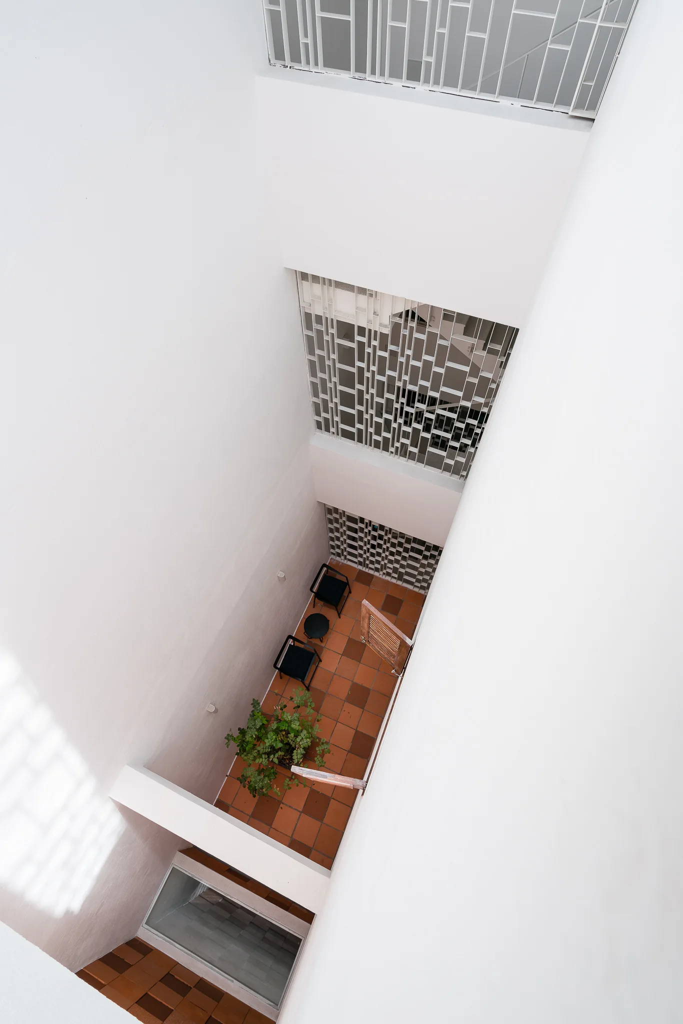 High-angle view of a white minimalist light well courtyard featuring orange terracotta floor tiles, black chairs, and geometric metal screens.