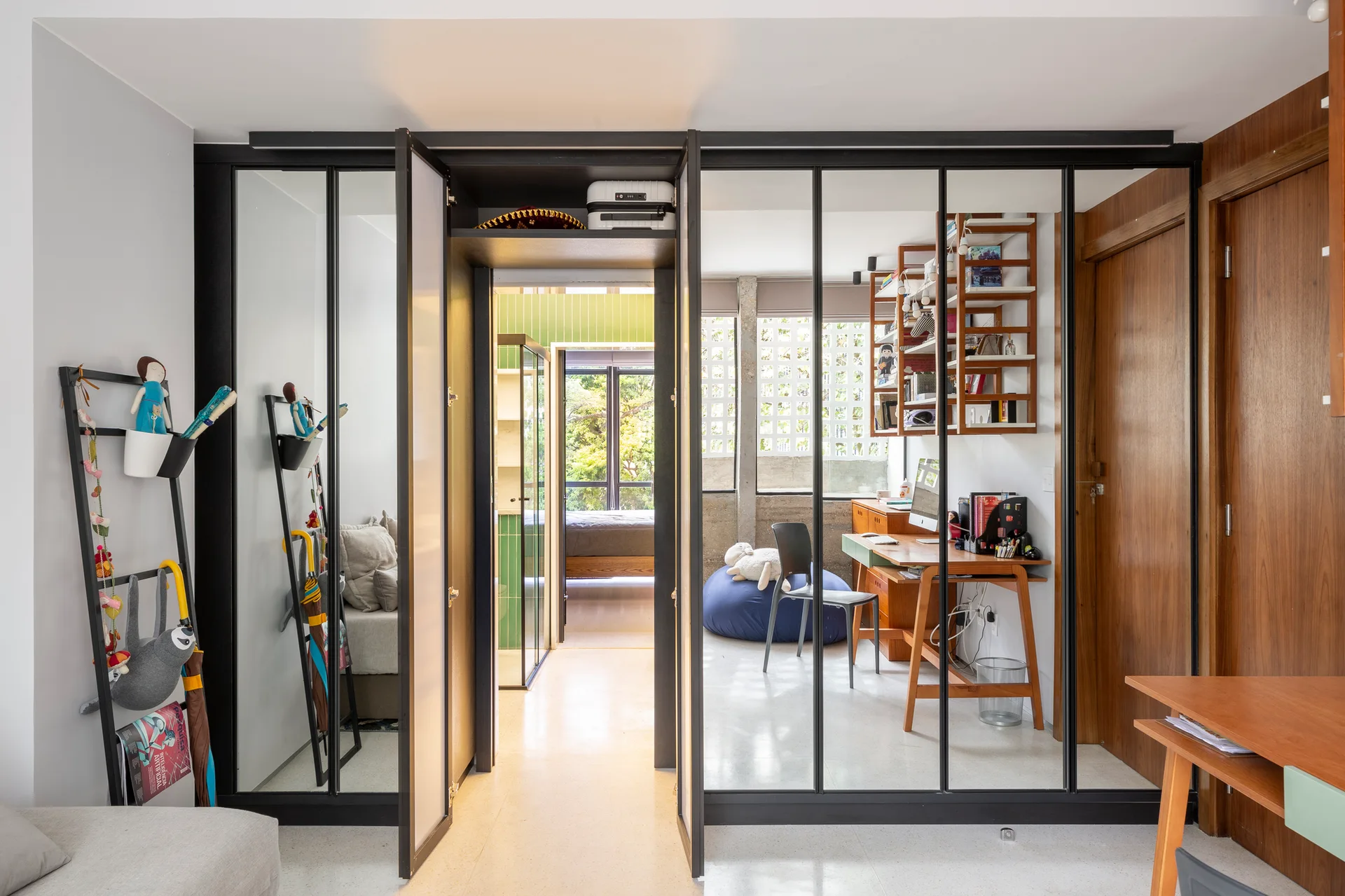 A modern room divided by a black metal-framed glass partition, showing a workspace with a wooden desk, office chair, and a blue bean bag.