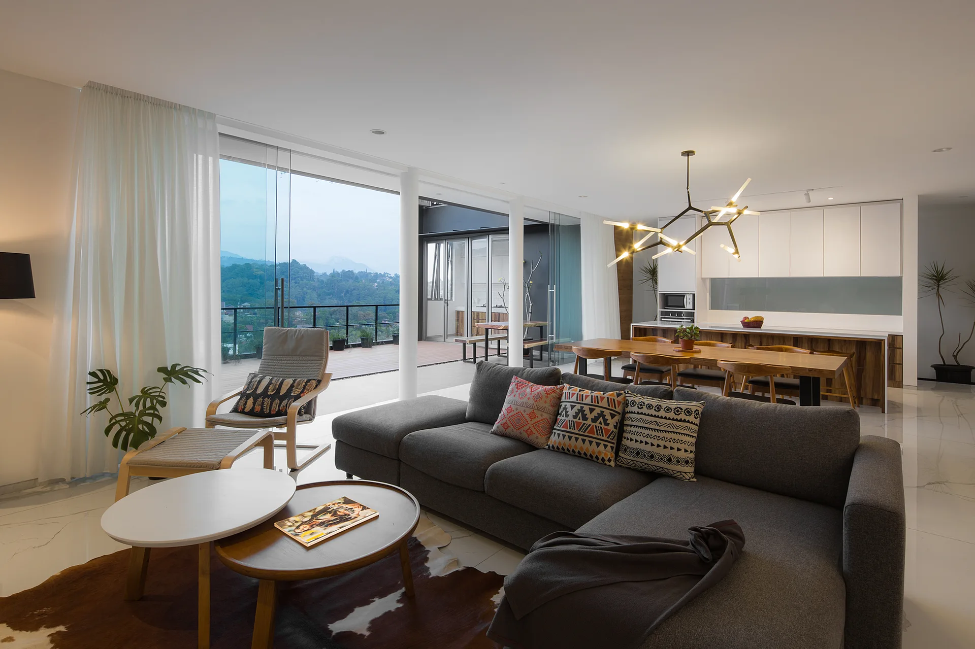 A modern open-plan living room features a grey sectional sofa, nesting coffee tables, and a wooden dining set next to large glass balcony doors.