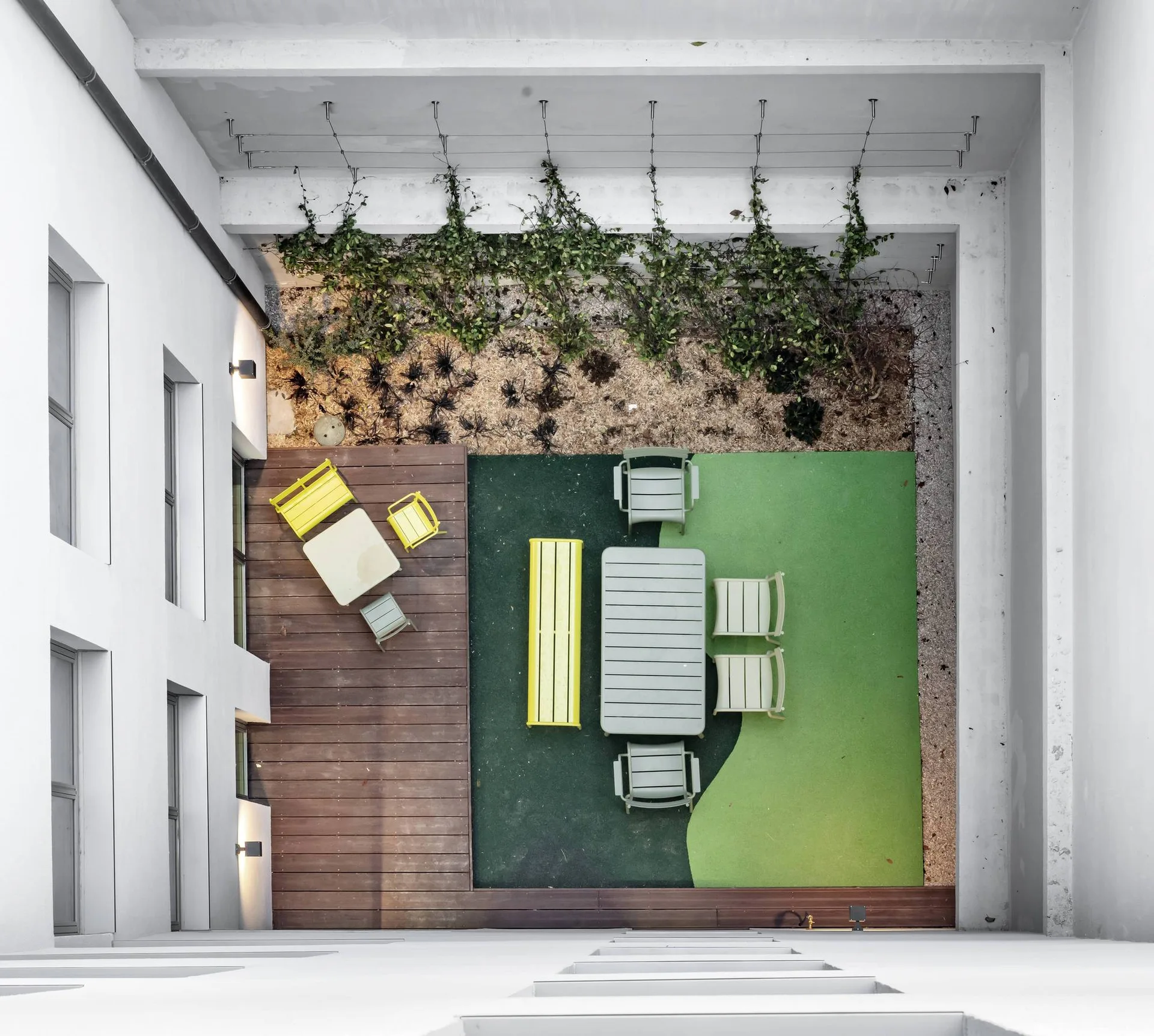 A top-down view of an enclosed courtyard with white walls, a wooden deck, a green synthetic floor, and modern colorful outdoor dining furniture.