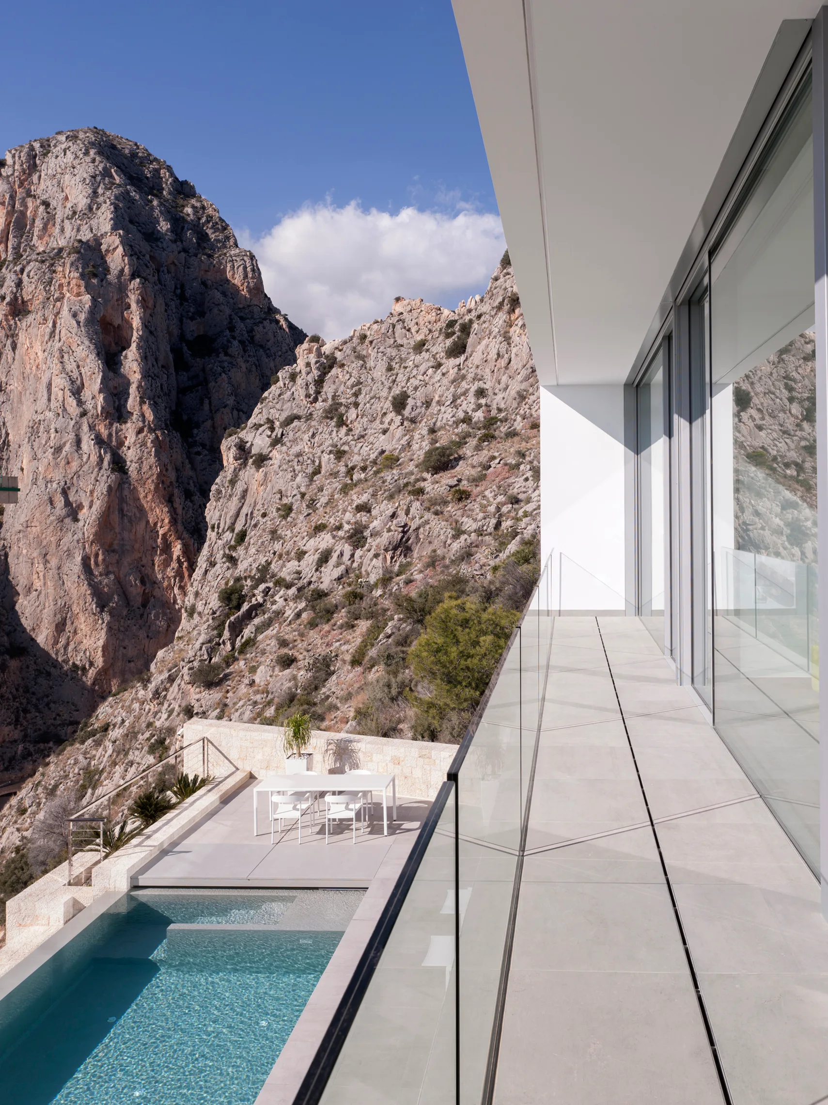 A modern grey-tiled terrace with a frameless glass railing overlooks a swimming pool, outdoor dining table, and rugged limestone mountains.