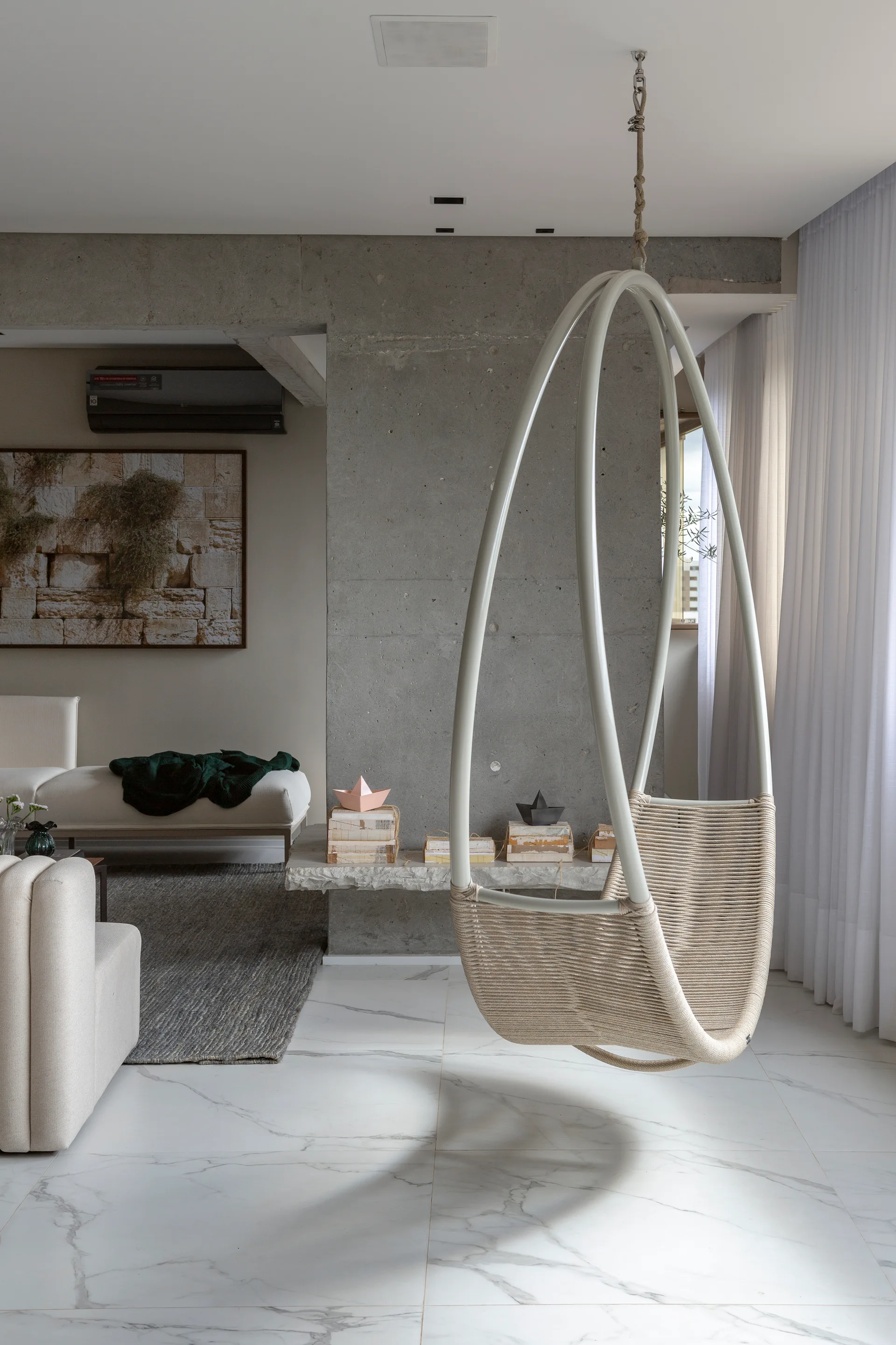 A woven hanging chair suspends from the ceiling of a modern living room featuring an exposed concrete wall and polished white marble flooring.
