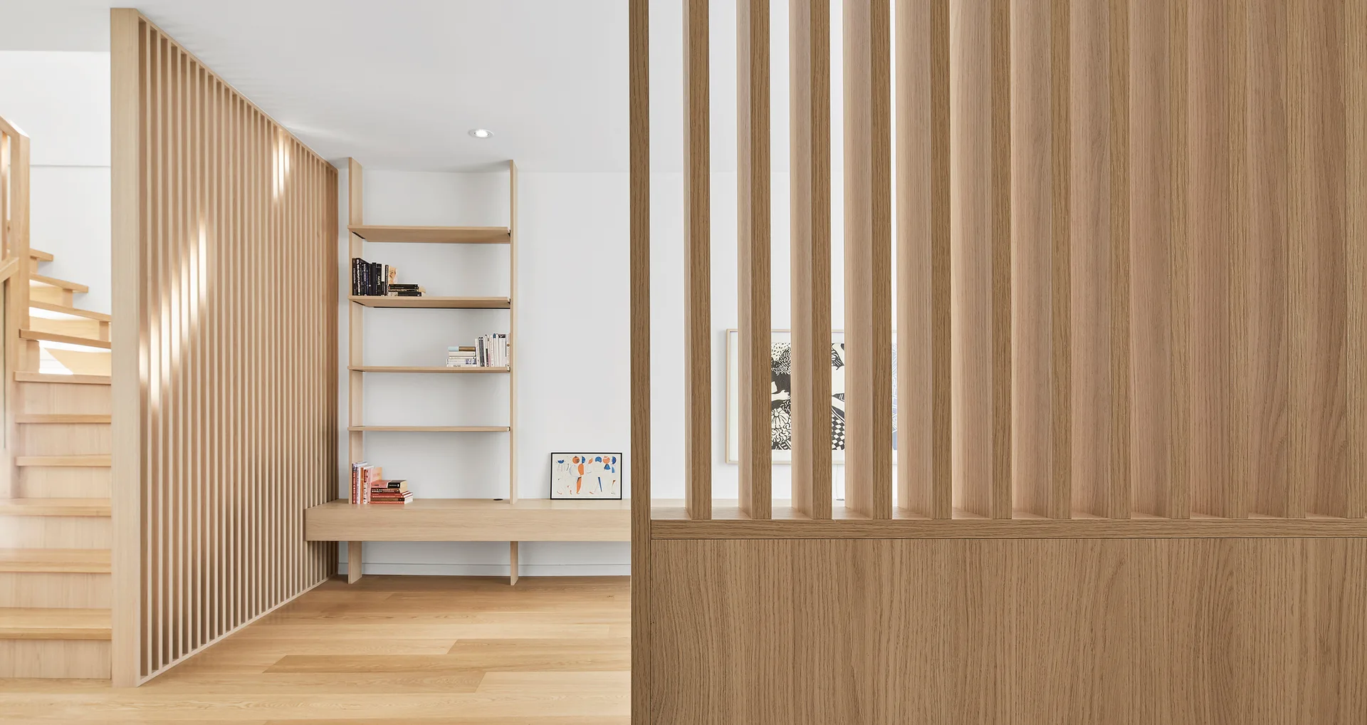 A minimalist hallway featuring vertical light wood slat room dividers, matching hardwood flooring, a wooden staircase, and a built-in shelving unit.