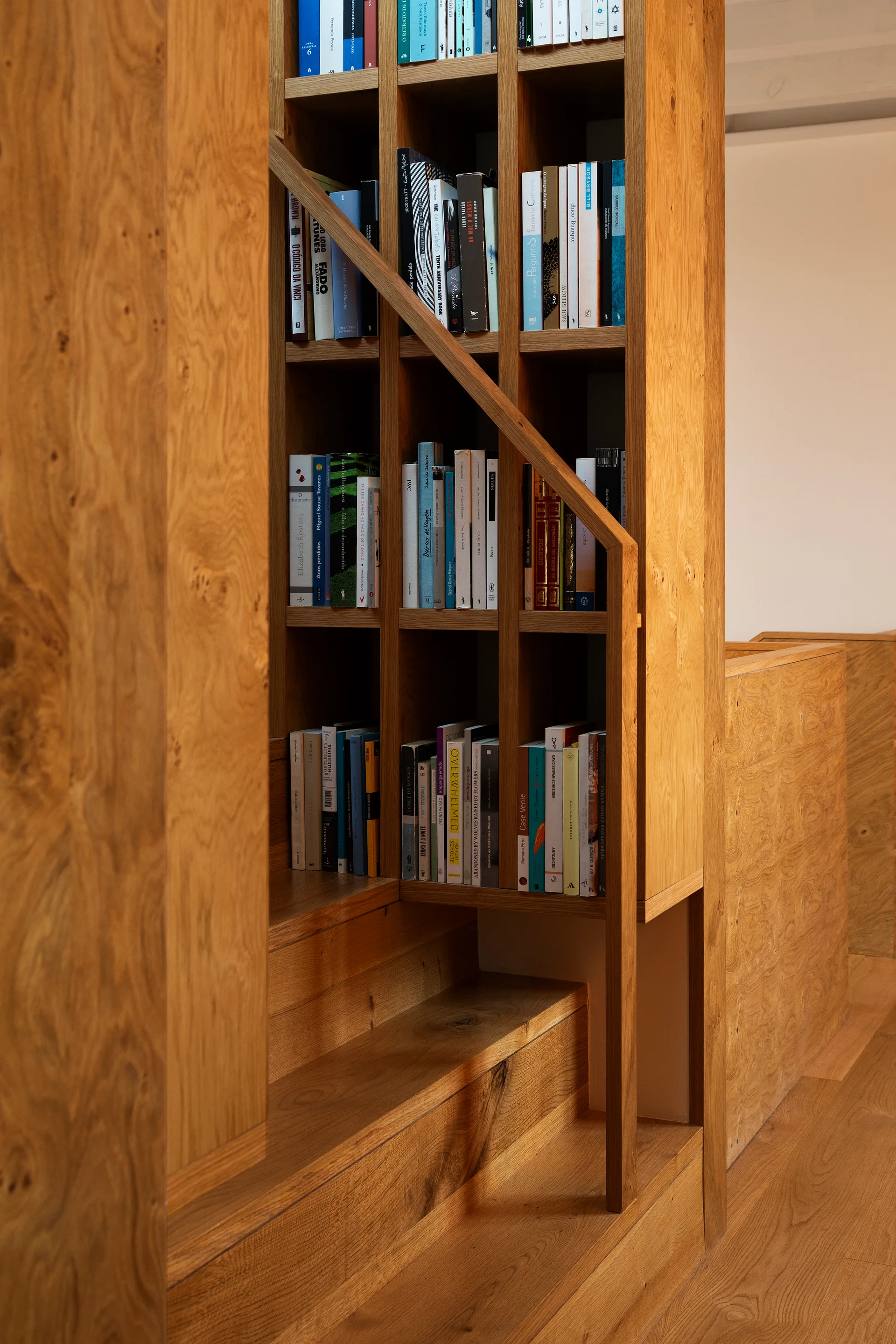 A close-up of a warm wood staircase featuring integrated bookshelves filled with colorful books and a matching diagonal handrail.