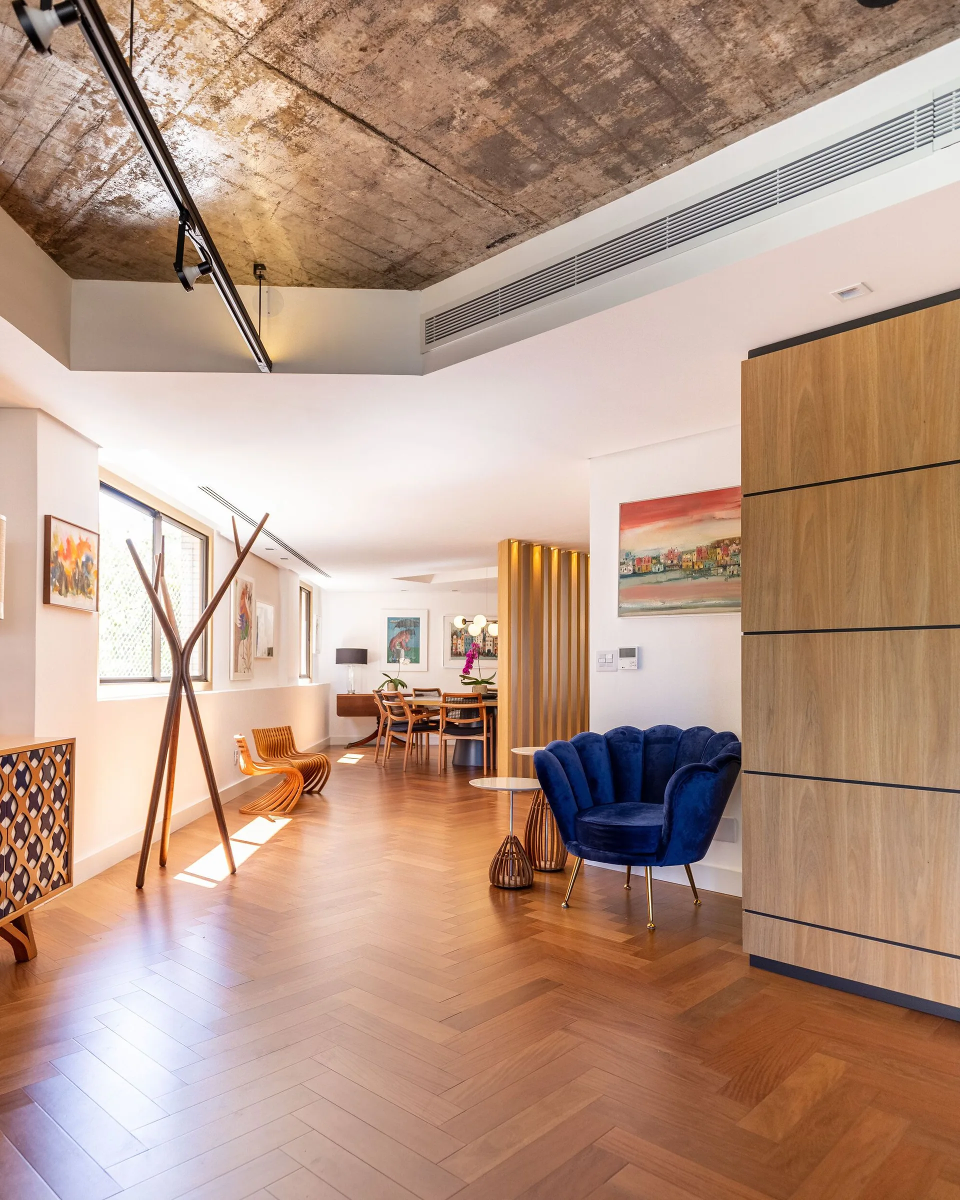 A modern open-concept room features an exposed concrete ceiling, warm herringbone hardwood floors, a blue velvet scalloped armchair, and a dining area.