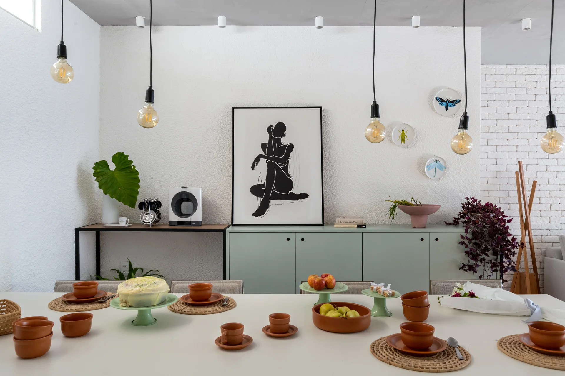 A white dining table styled with terracotta tableware and woven placemats stands before a matte mint green sideboard and a textured plaster wall.