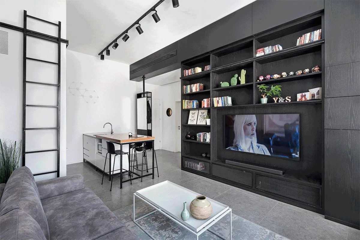A modern living room features a large black built-in wall unit with a television, a grey sofa, a rolling ladder, and track lighting.