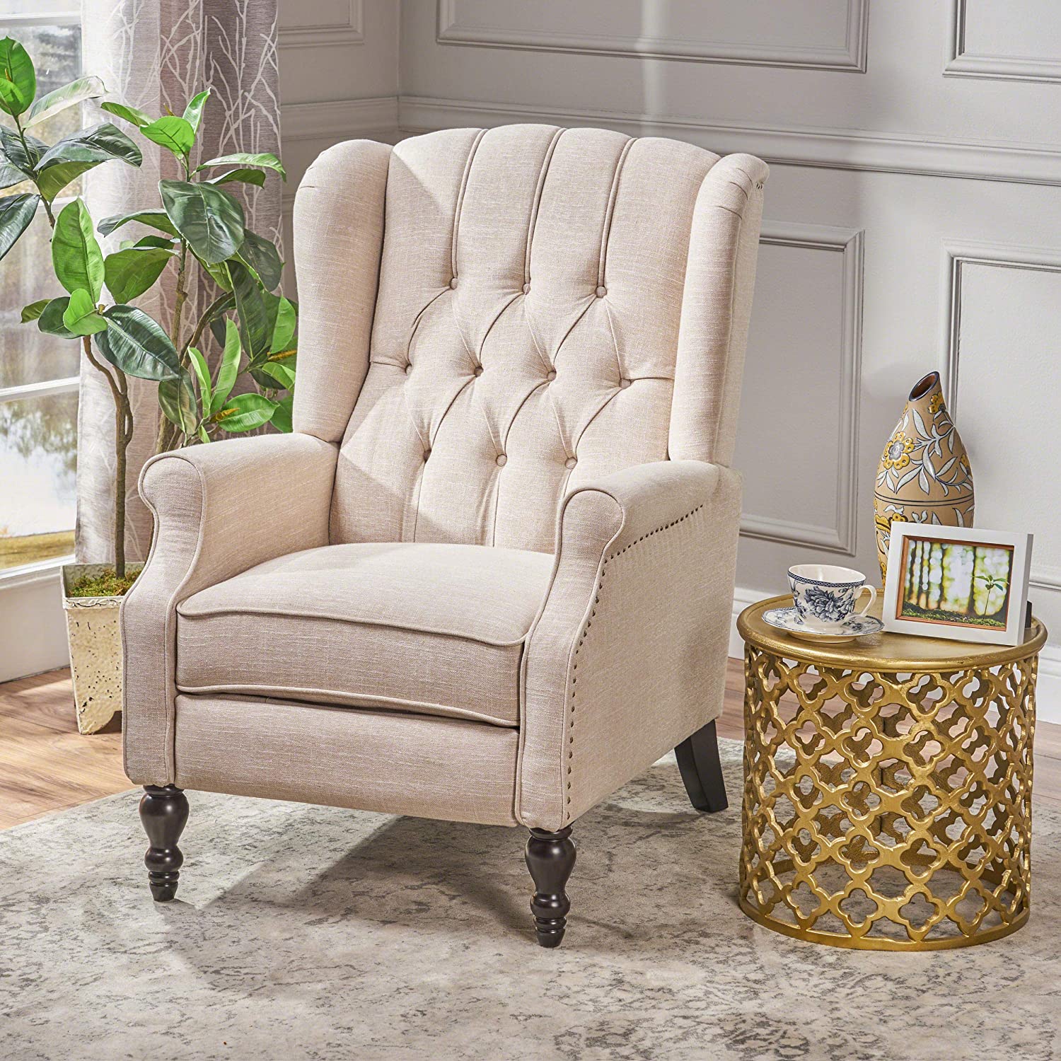 A cream tufted wingback armchair sits on a patterned rug next to a round gold metal cutout side table decorated with a teacup.