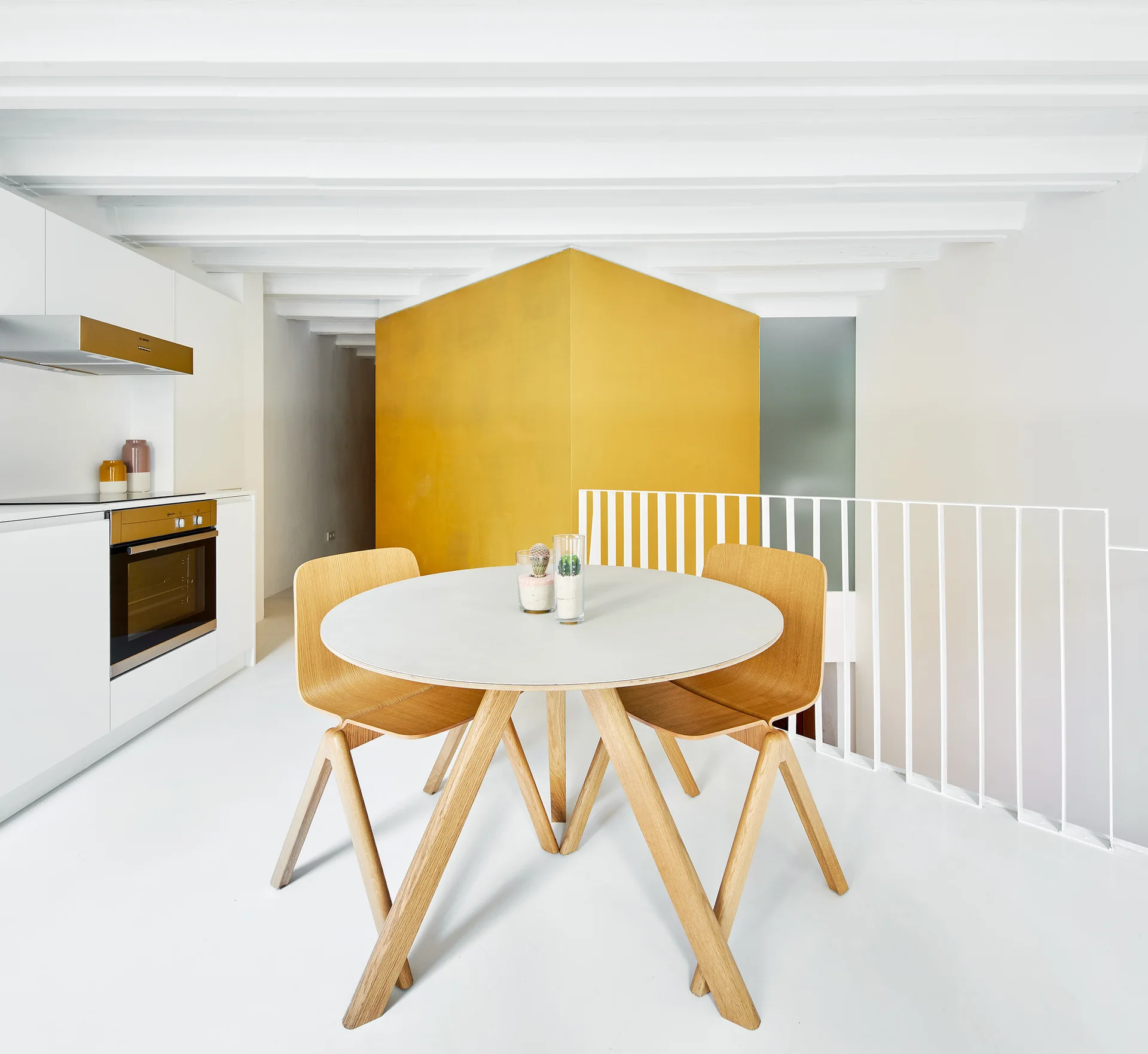 A round white dining table with light wood chairs sits in a minimalist room featuring a bold yellow architectural partition and white painted ceiling beams.