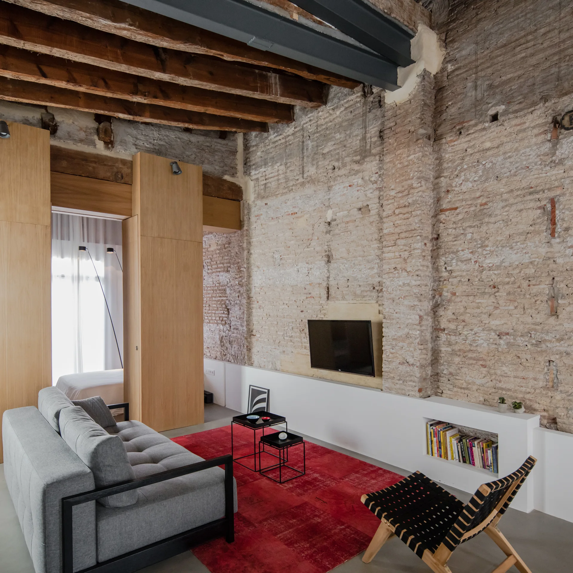 A modern loft living room features a grey sofa, a vibrant red rug, an exposed brick wall, and rustic wood ceiling beams.