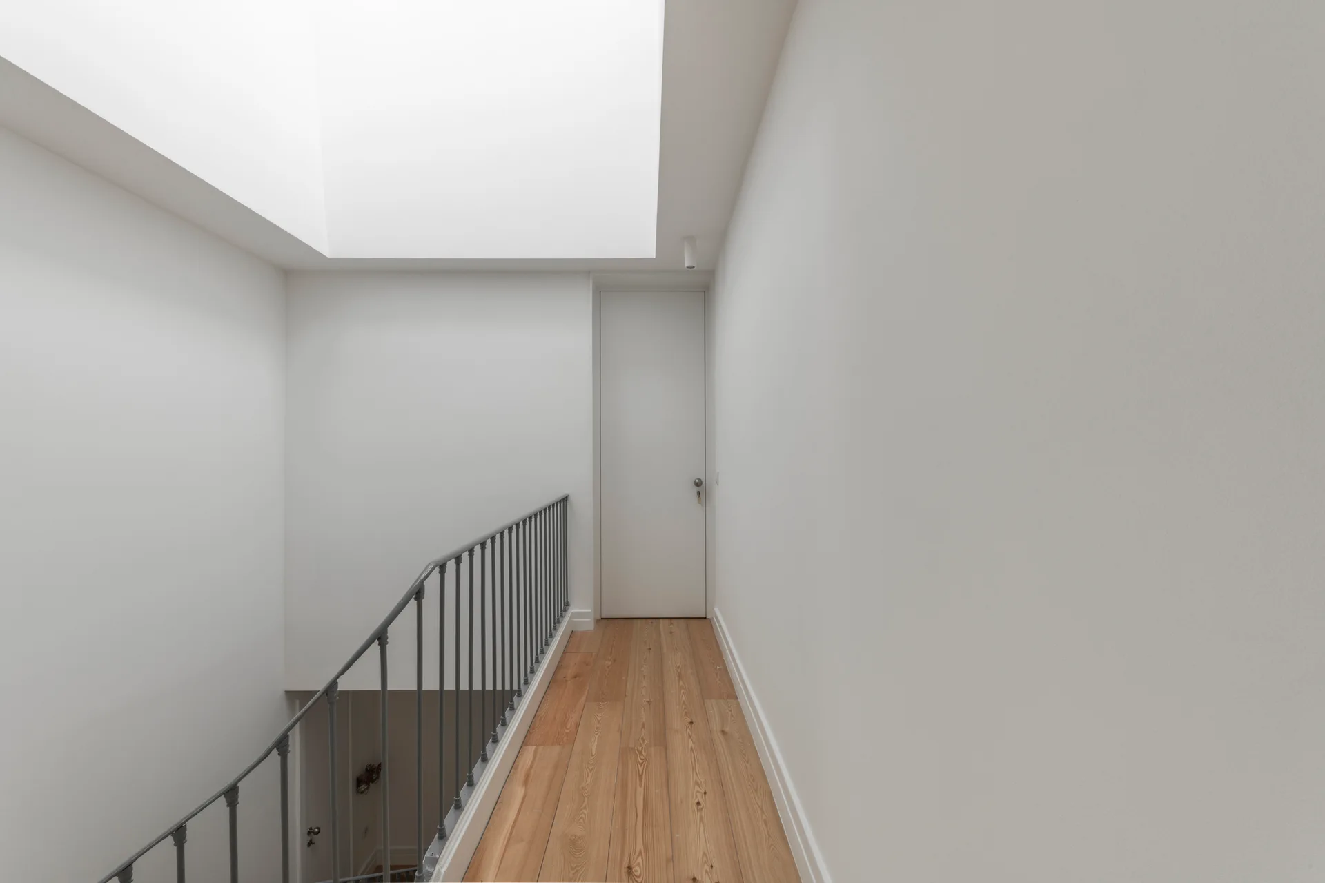 A narrow white hallway features natural wood floors, a metal stair railing, a closed door at the end, and a bright overhead skylight.