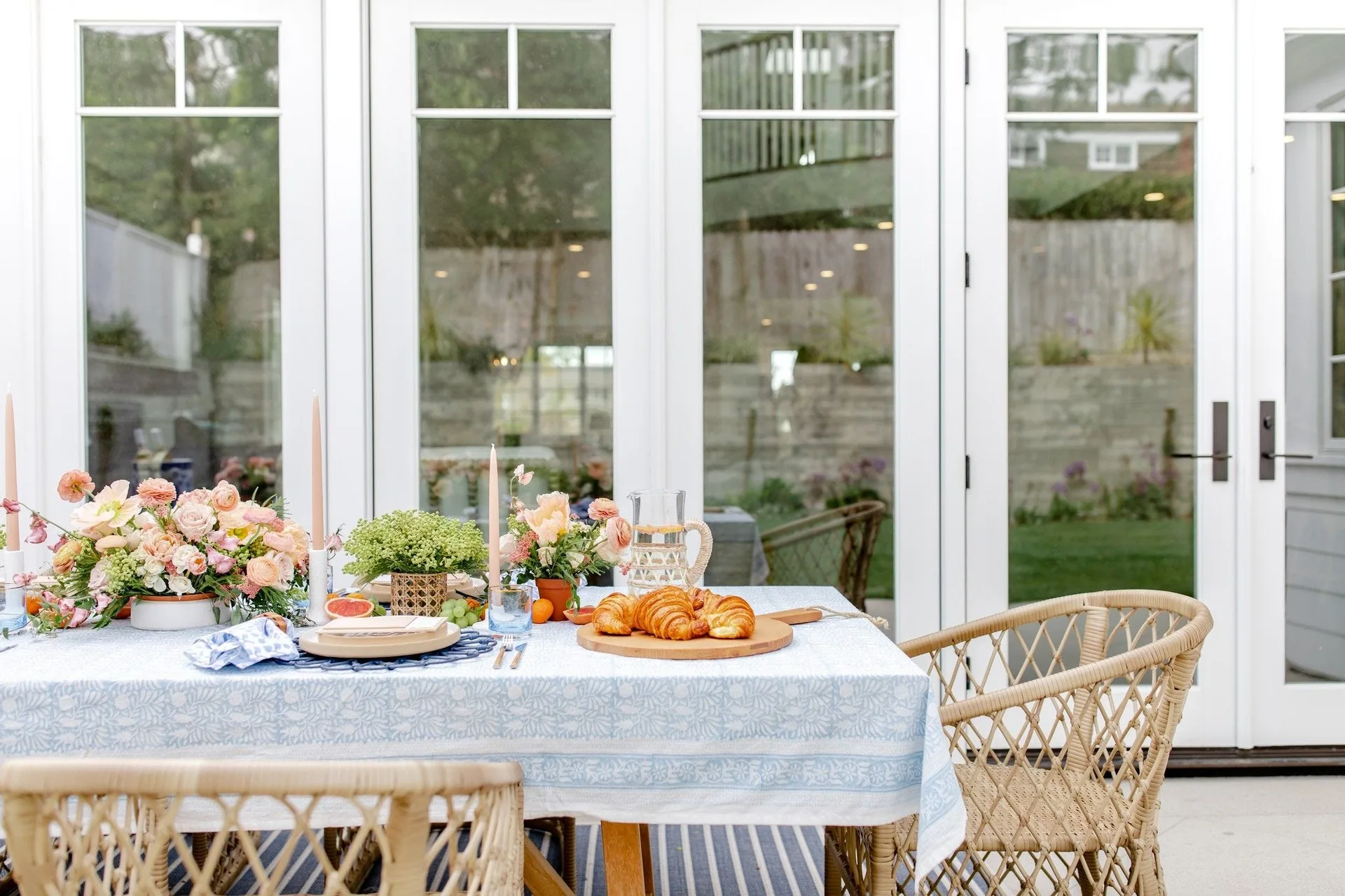 A dining table set with a blue patterned tablecloth, floral arrangements, and wicker chairs sits in front of white French doors.