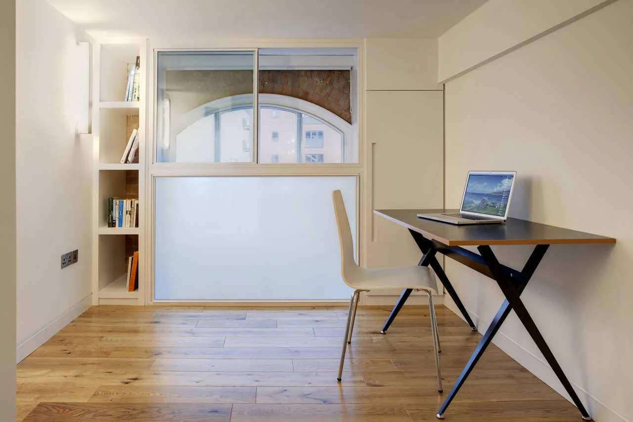 A minimalist home office features a black desk, a white chair, light wood floors, and built-in shelving next to a frosted window.