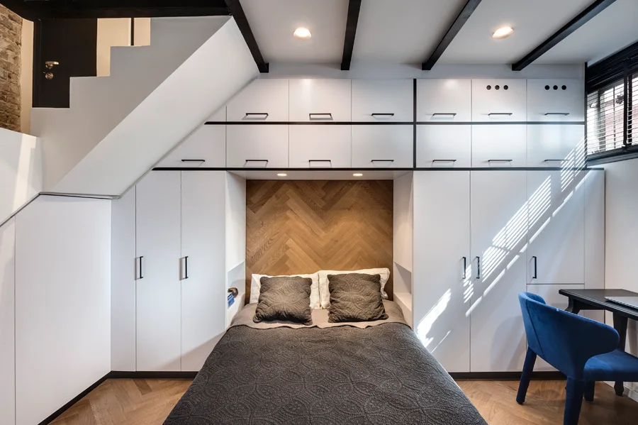 A compact bedroom features white built-in storage closets surrounding a bed with a warm herringbone wood panel headboard and dark gray bedding.
