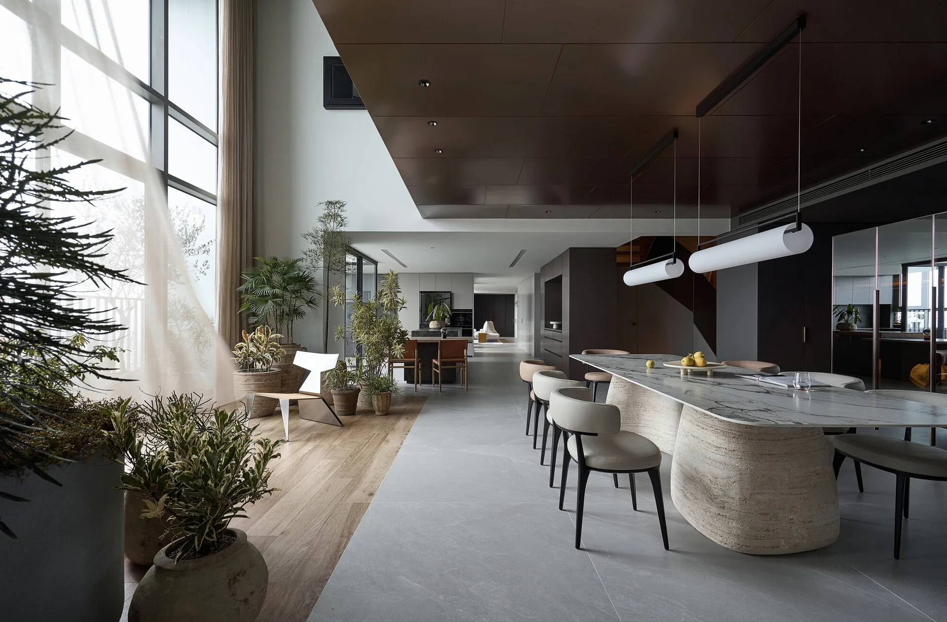 A modern dining room features a marble-topped table with sculptural stone bases, a dark paneled ceiling, and large potted plants by tall windows.