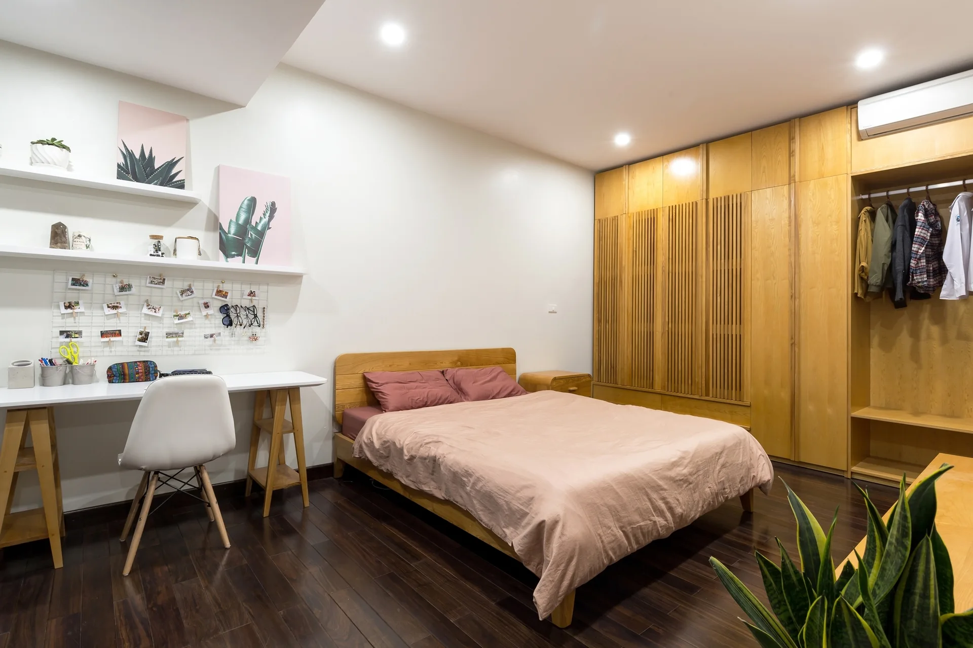 A modern bedroom features a wooden bed with pink linens next to a large slatted wood wardrobe and a white study desk.