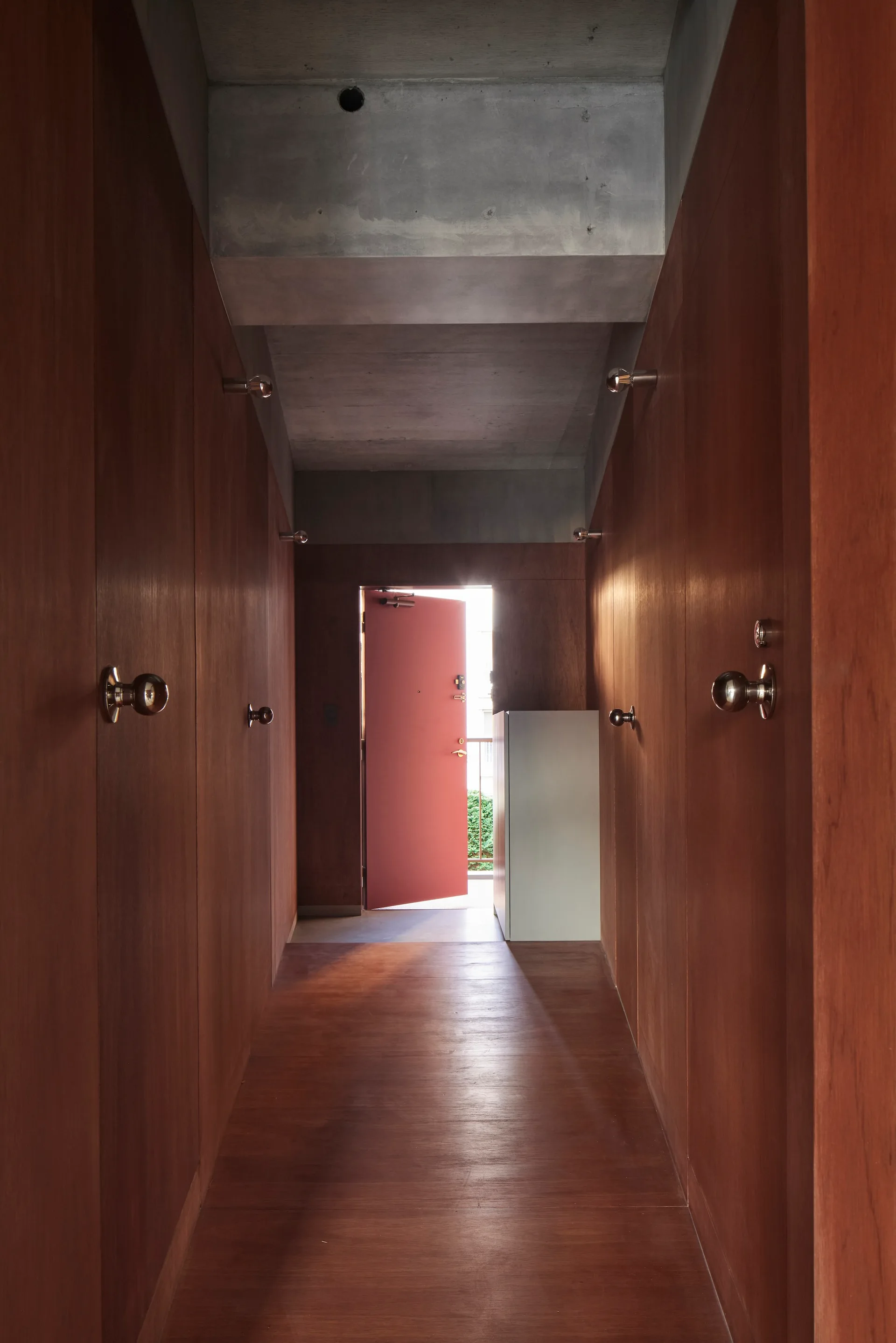 A narrow hallway lined with rich brown wood paneling and matching floors features an exposed concrete ceiling and an open red door at the far end.