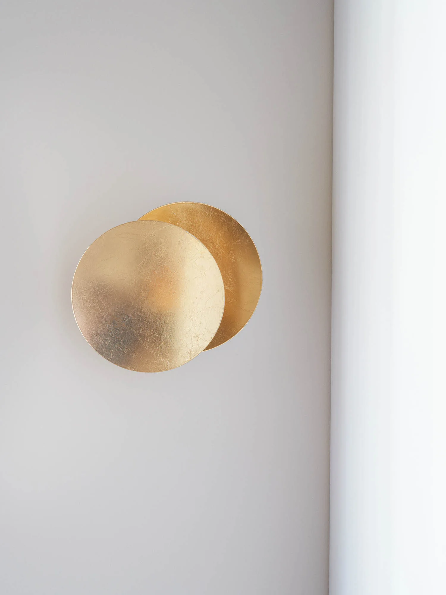 A modern circular gold metal wall sconce consisting of two overlapping discs is mounted on a smooth light grey wall.