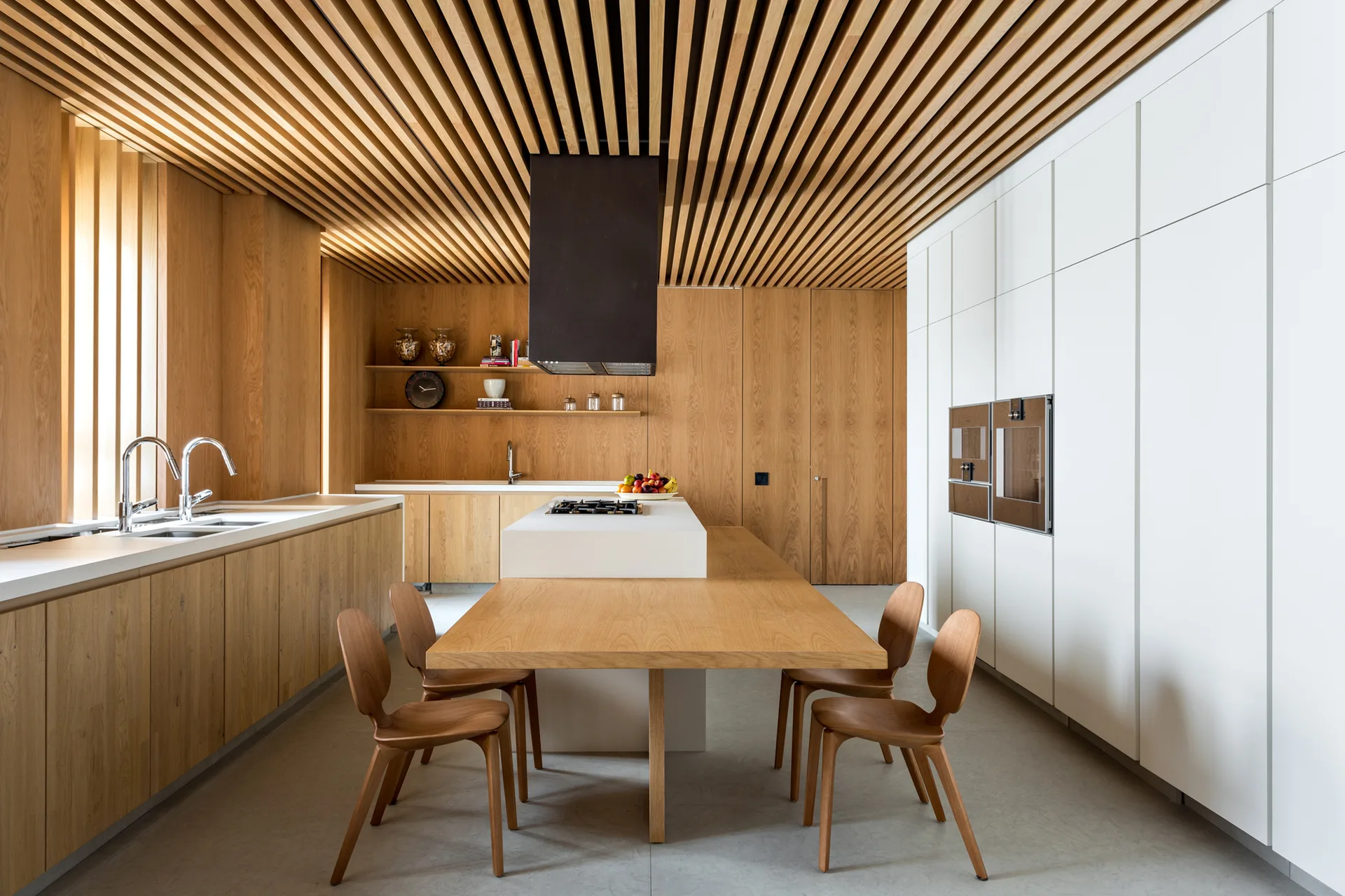 A modern kitchen featuring a natural wood slat ceiling, a white island with an attached oak table, and tall white cabinets.