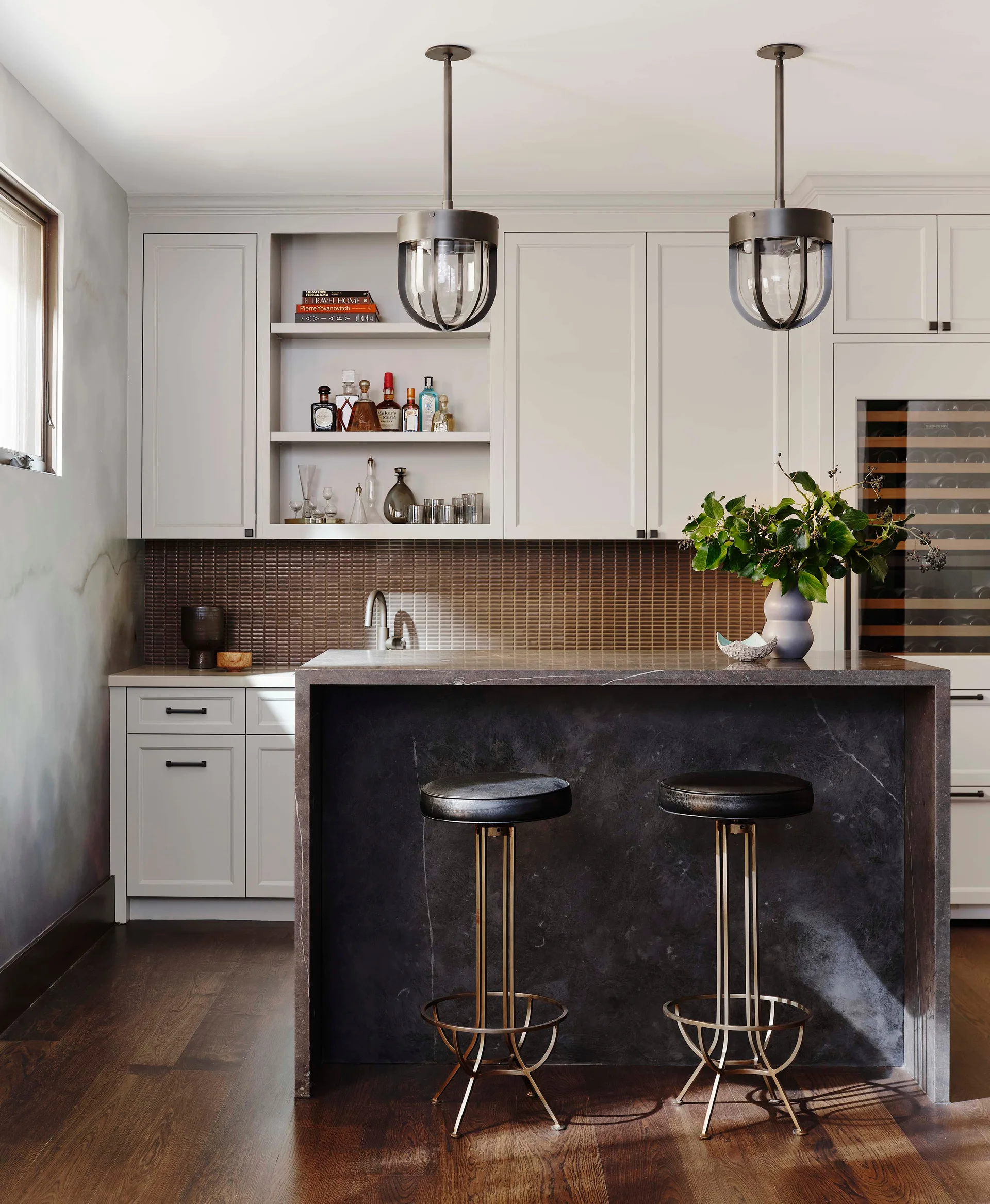A home bar featuring a dark stone waterfall island, two black and brass bar stools, light grey cabinets, and a textured tile backsplash.