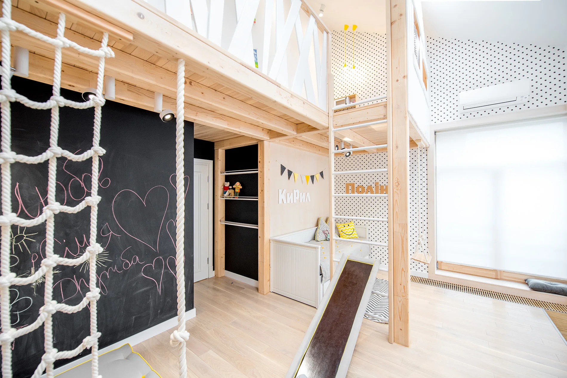 A children's bedroom features a natural wood loft bed with an integrated slide, a white daybed, and a large black chalkboard wall.