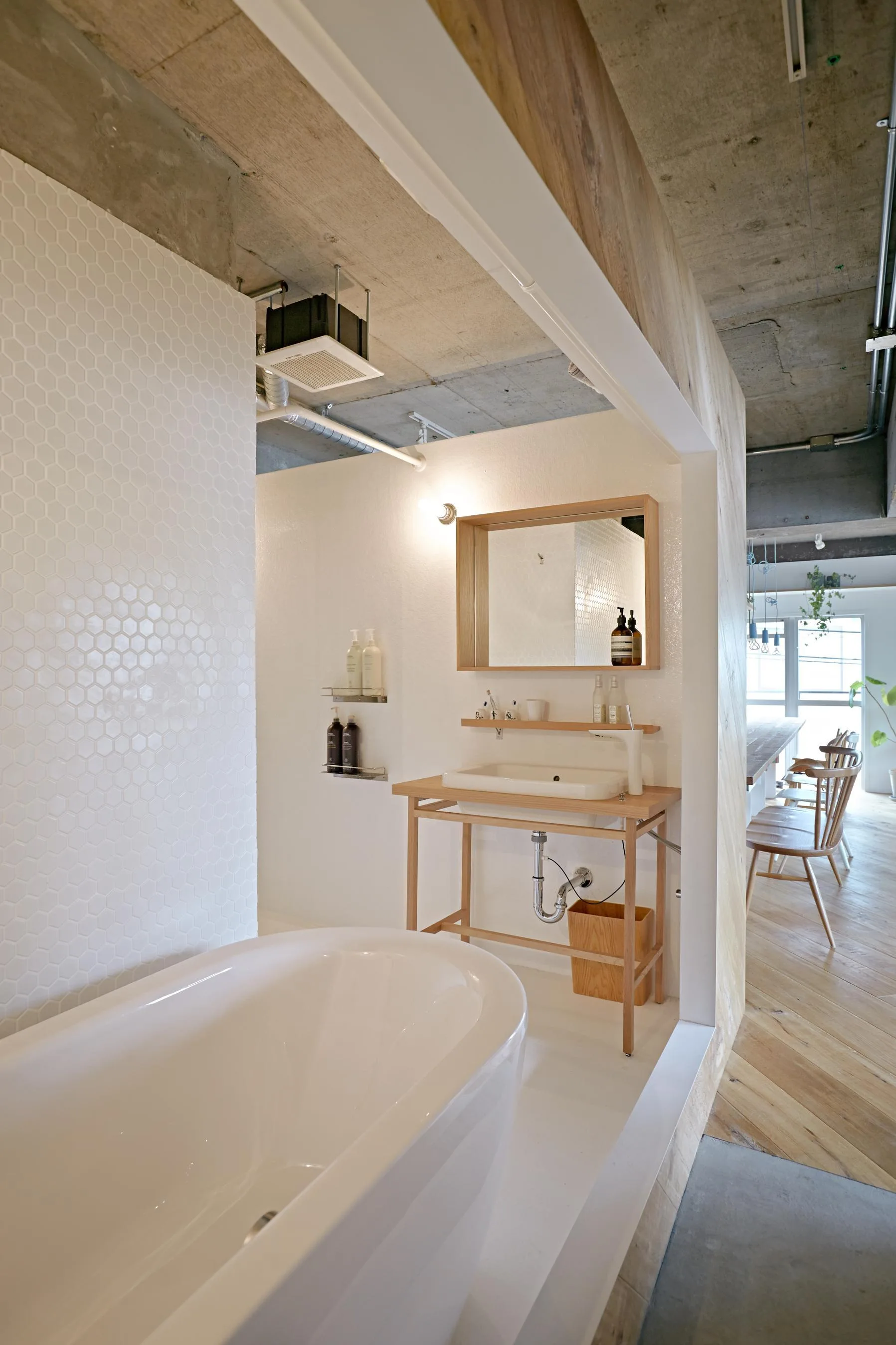 A minimalist bathroom features a white freestanding bathtub and open wood vanity under an exposed concrete ceiling next to a dining area.
