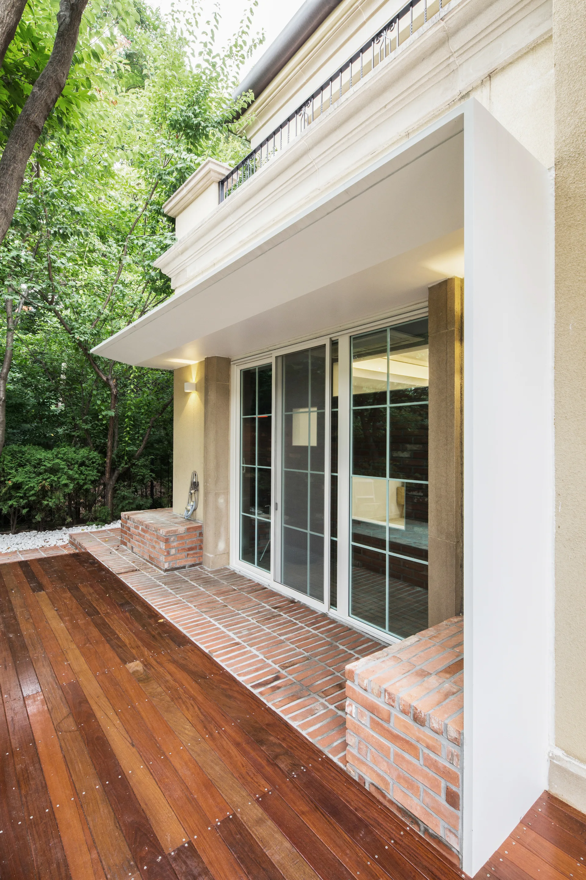 An outdoor patio featuring rich wood plank decking, a red brick border, and sliding glass doors under a modern white canopy frame.