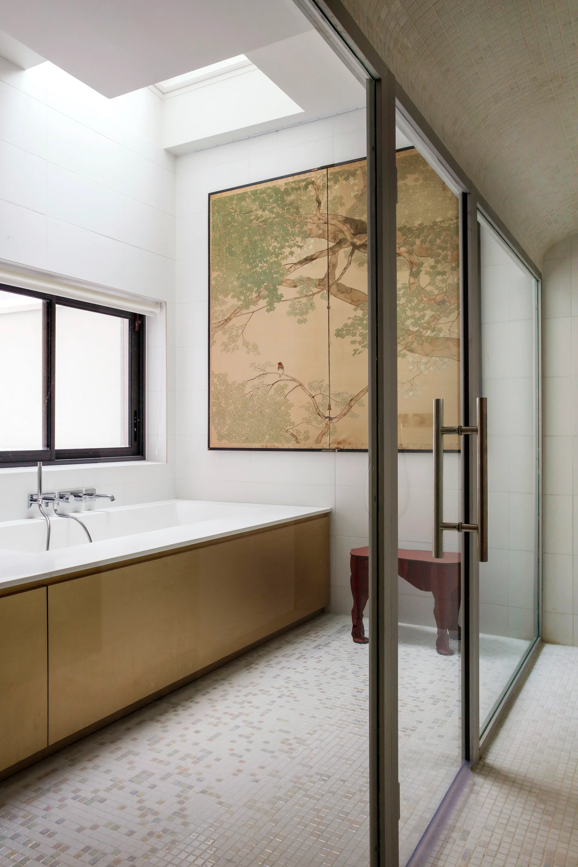 A modern white bathroom featuring a rectangular bathtub with a gold base, a glass shower, mosaic tile floors, and a large nature painting under a skylight.