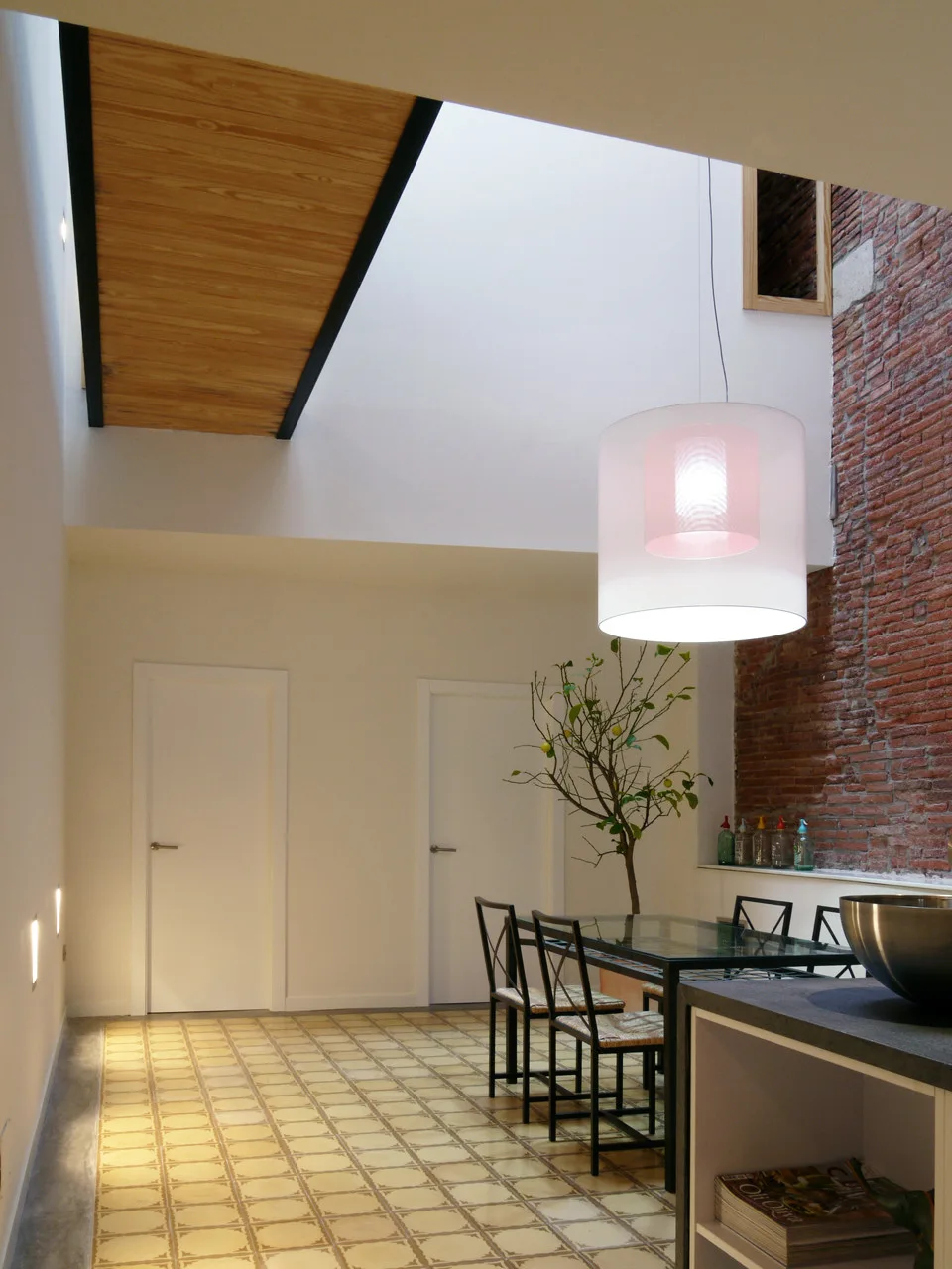 A contemporary dining room featuring a glass table, black chairs, a large drum pendant light, an exposed brick wall, and yellow patterned floor tiles.