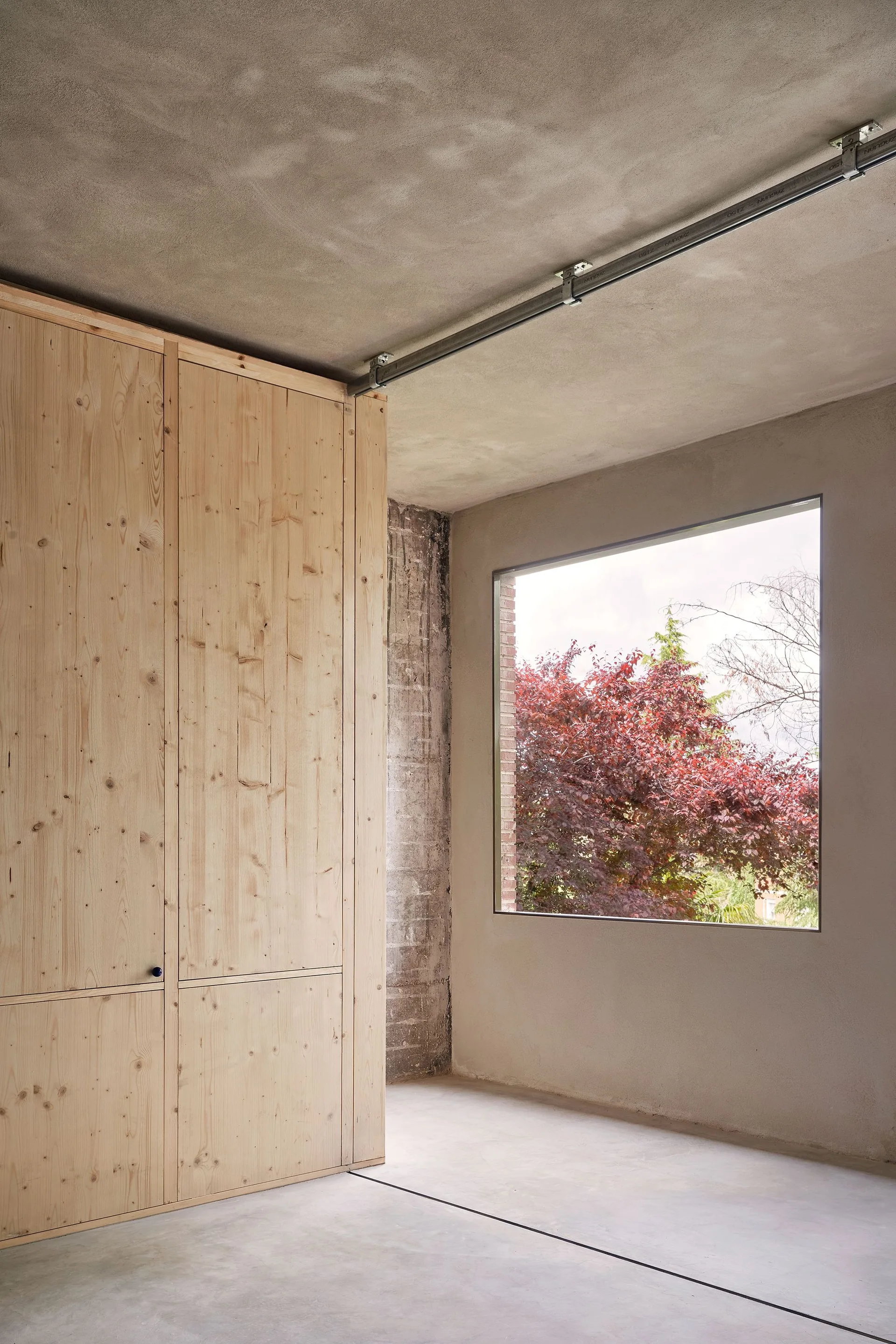 A minimalist room features raw concrete walls, a smooth grey floor, floor-to-ceiling light wood cabinets, and a square window showcasing trees outside.