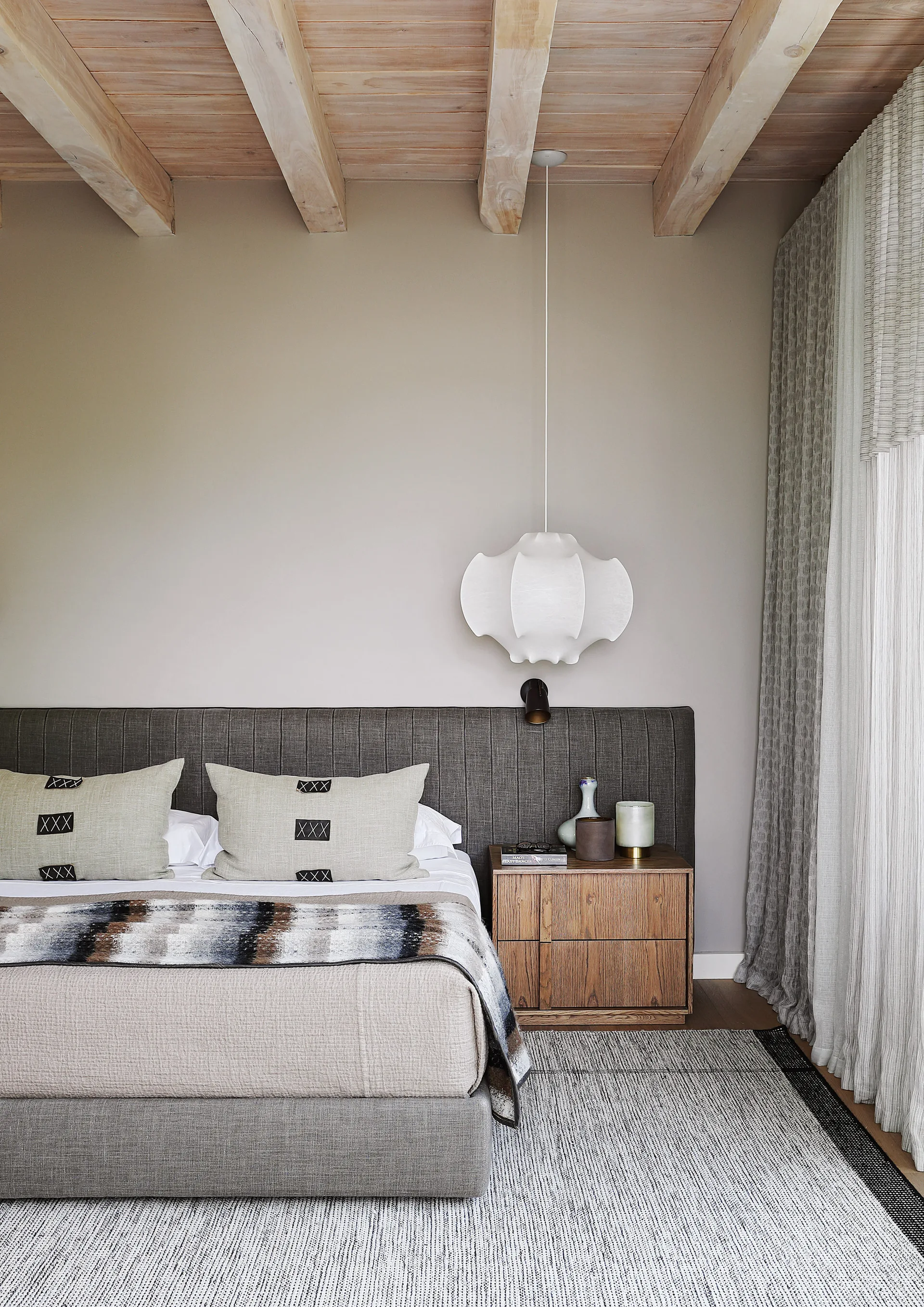 A modern bedroom features a low-profile bed with a gray upholstered headboard, a wooden nightstand, and exposed natural wood beams on the ceiling.