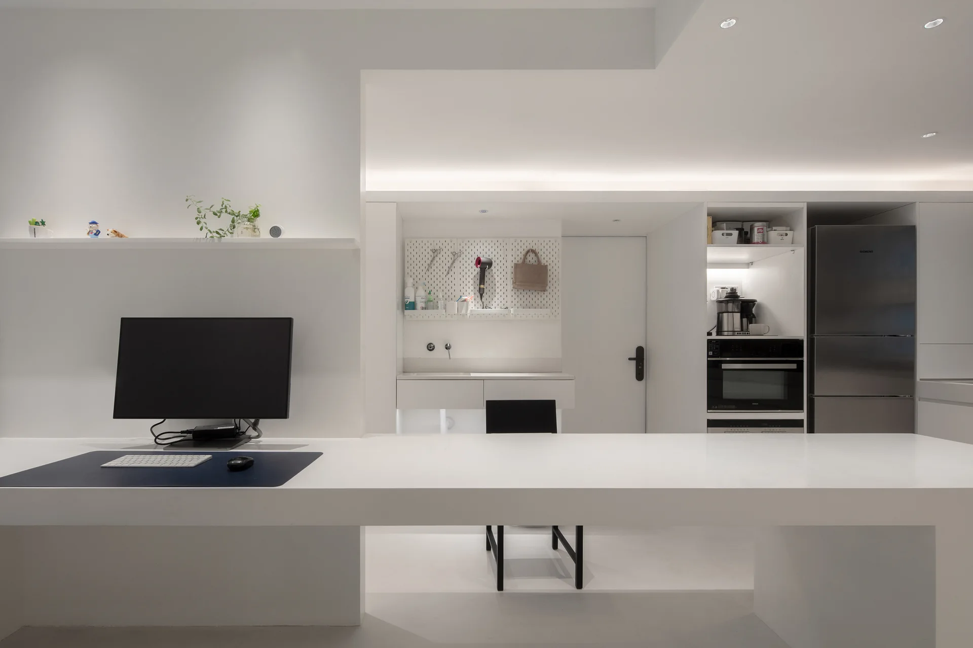 A minimalist all-white kitchen features a long integrated desk with a computer monitor, built-in stainless steel appliances, and a wall pegboard.