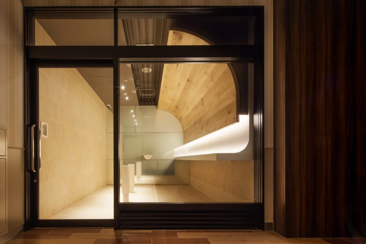 A modern entryway seen through a black-framed glass door, featuring beige tiled walls, a built-in shelf, and a large curved wood wall detail.