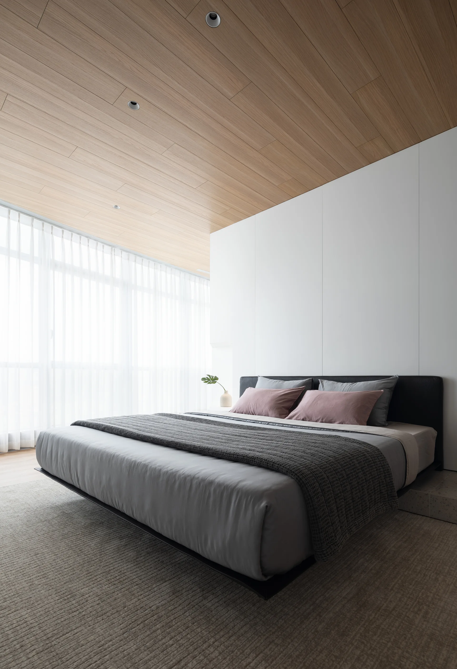 A minimalist bedroom features a low-profile platform bed with grey linens, white paneled walls, and a warm wood plank ceiling with recessed lighting.