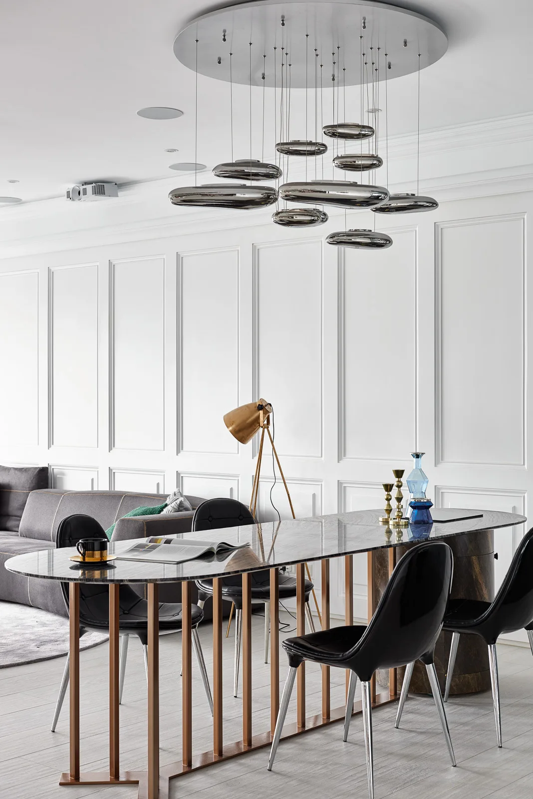 A contemporary dining room features an oval marble-topped table, glossy black chairs, and a chrome pebble chandelier against a white paneled wall.