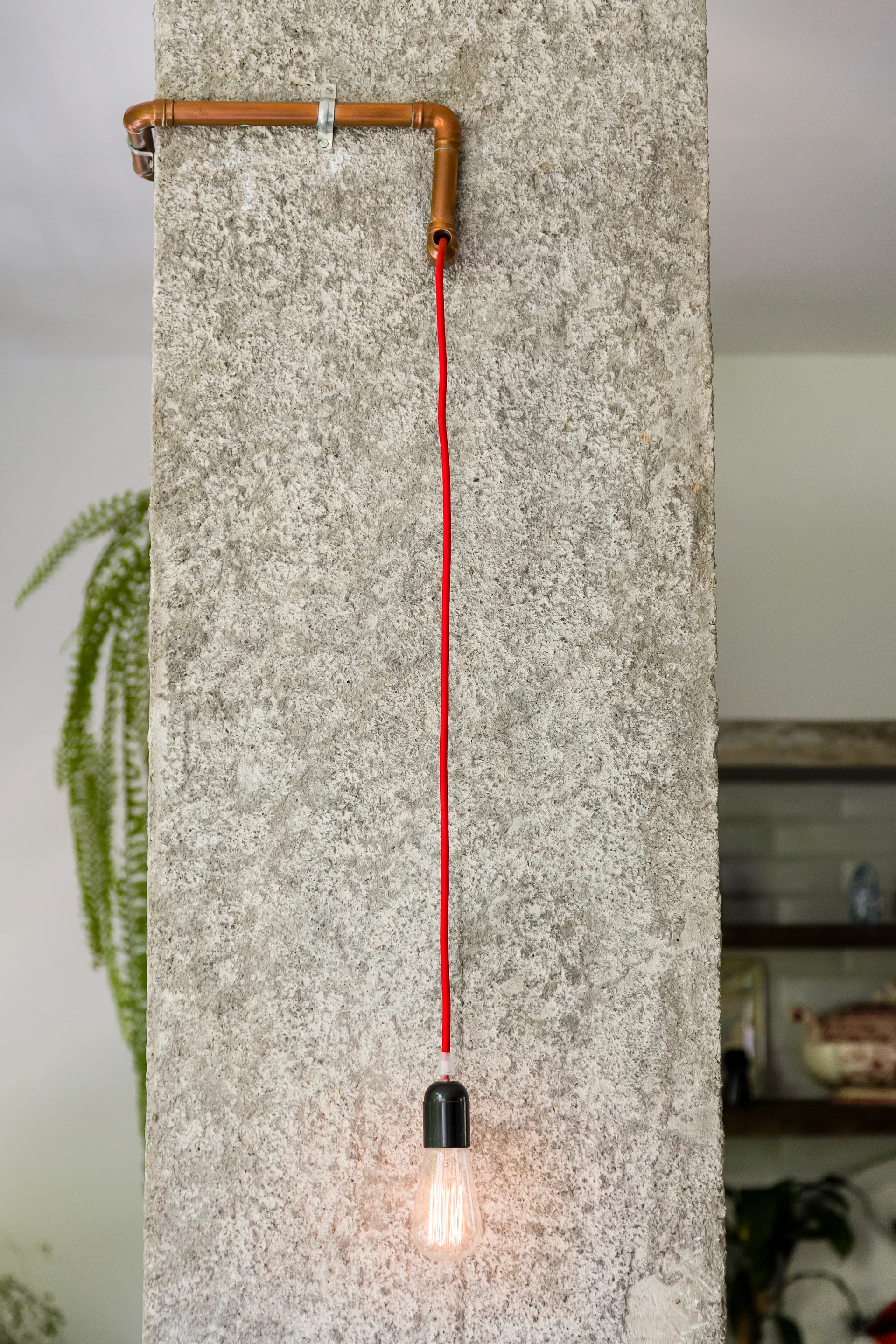 A bare light bulb hangs from a bright red cord connected to copper pipes mounted on a textured concrete pillar.
