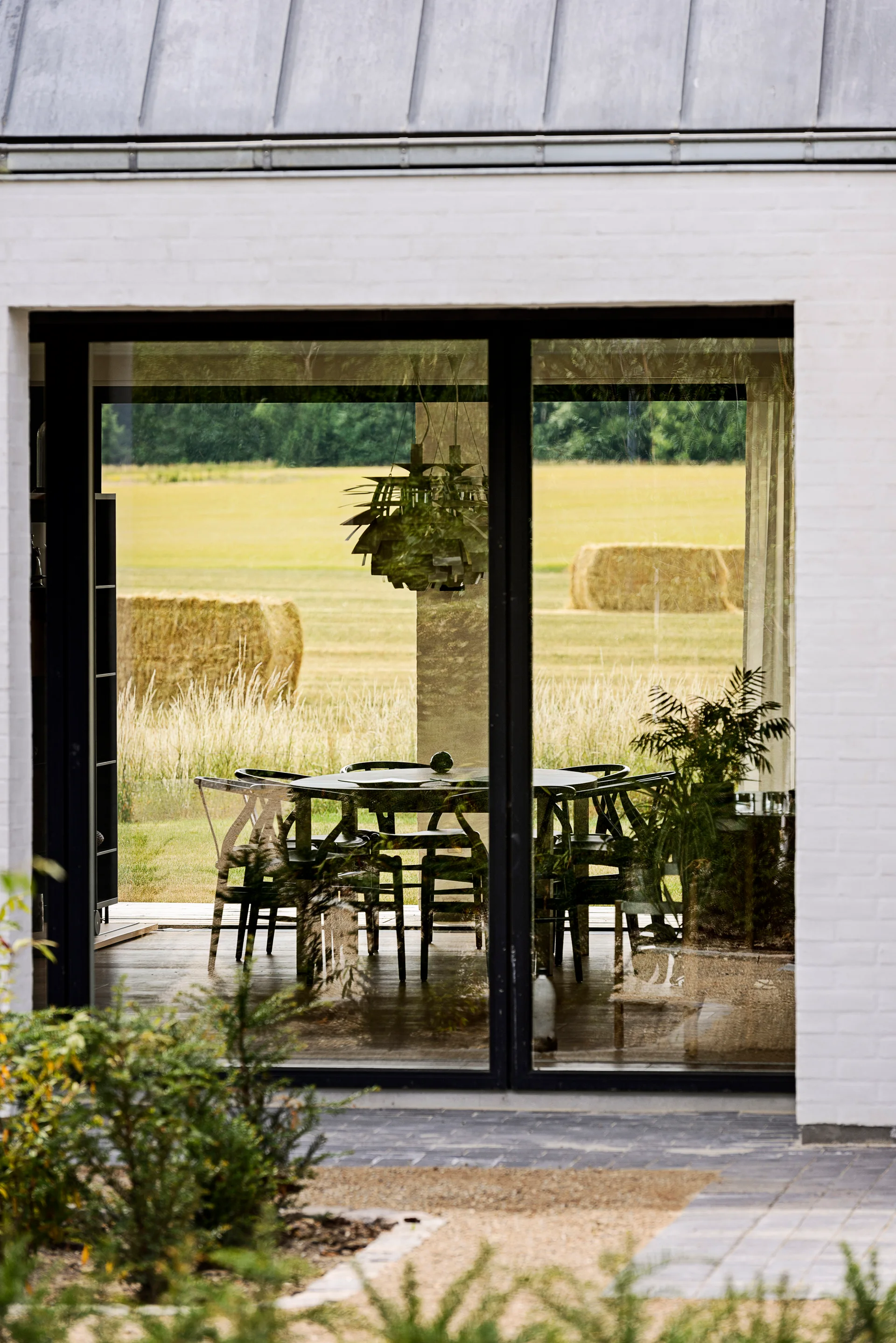 A view through a large window shows a round black dining table and chairs in a modern room, with a scenic field of hay bales in the background.