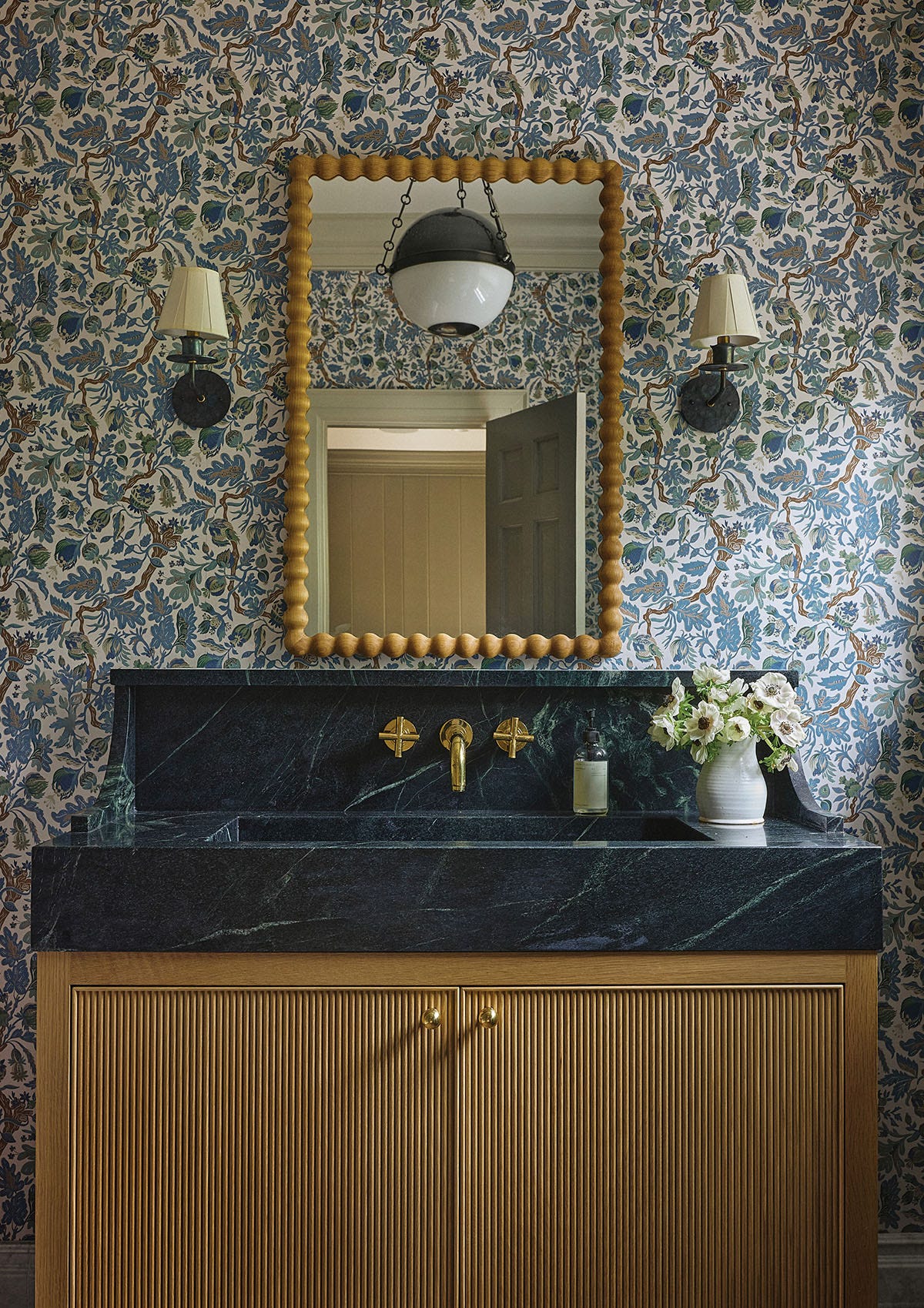 A powder room features a fluted wood vanity with a dark green marble countertop, wall-mounted brass fixtures, and blue botanical wallpaper.